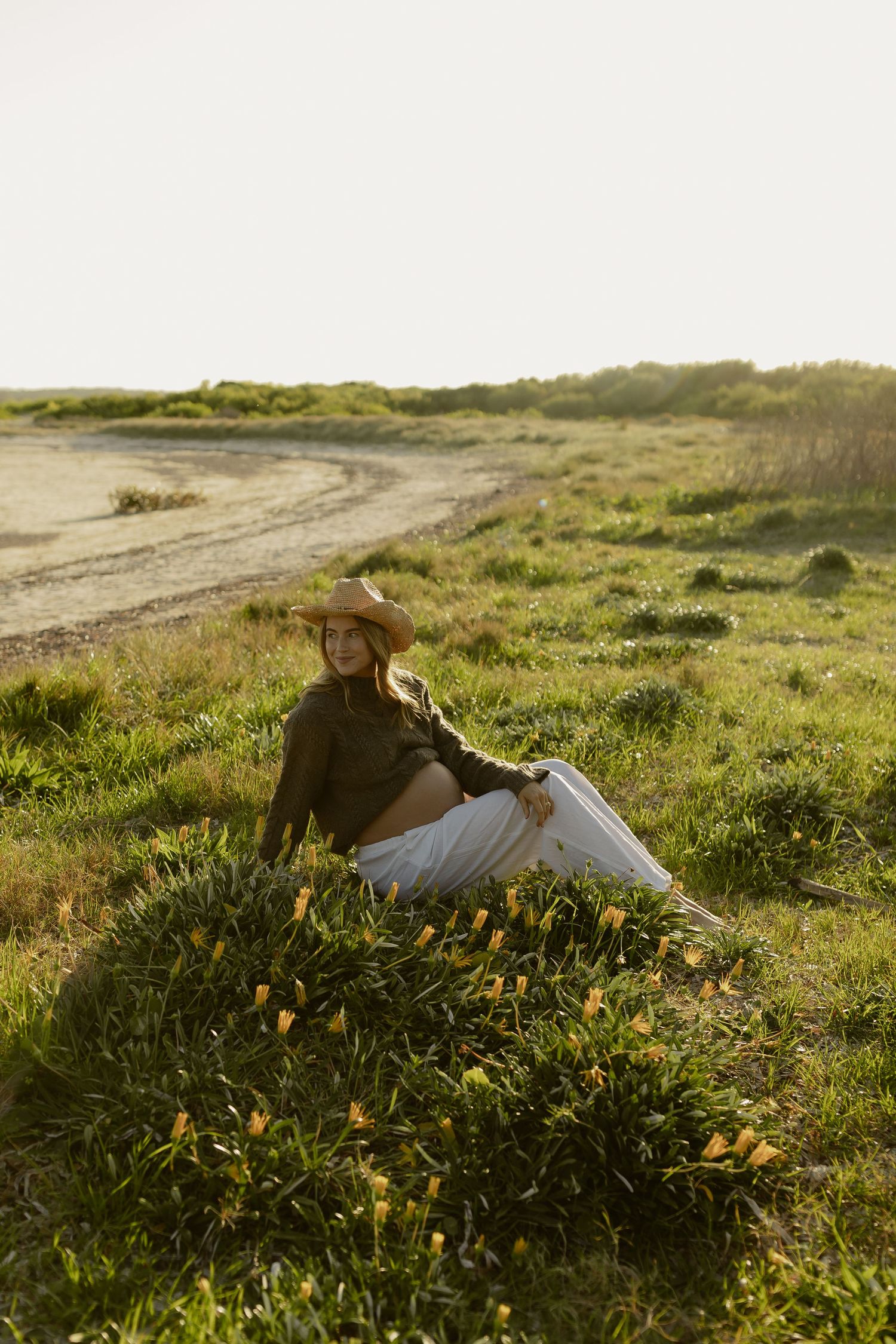 Woman in cowboy hat and white skirt sitting in green field with wildflowers at golden hour.