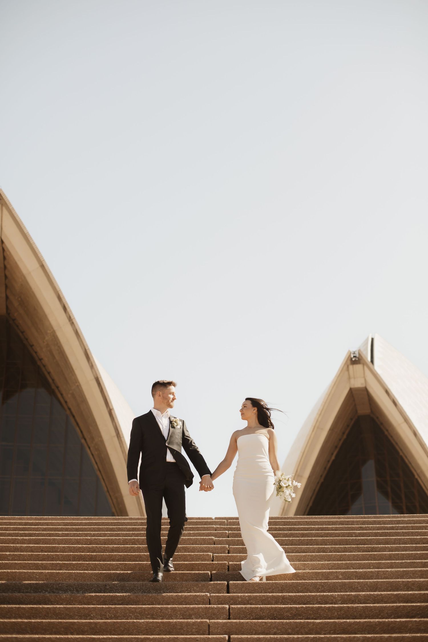 Wedding couple holds hands while walking down stairs at Sydney Opera House during golden hour.