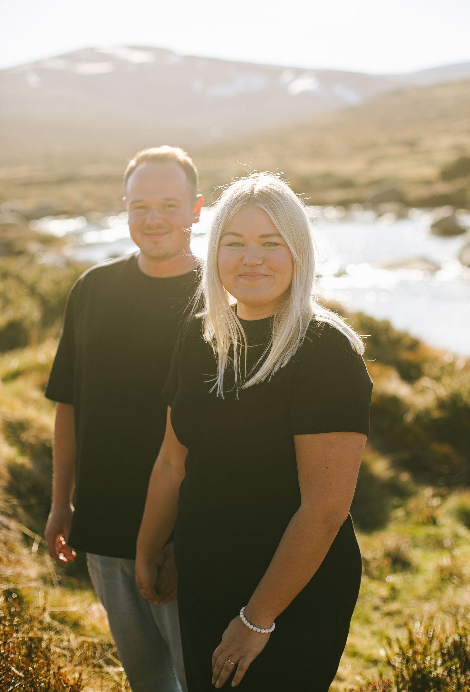 Two people in black shirts enjoy a sunny day outdoors with mountains and water in the background.