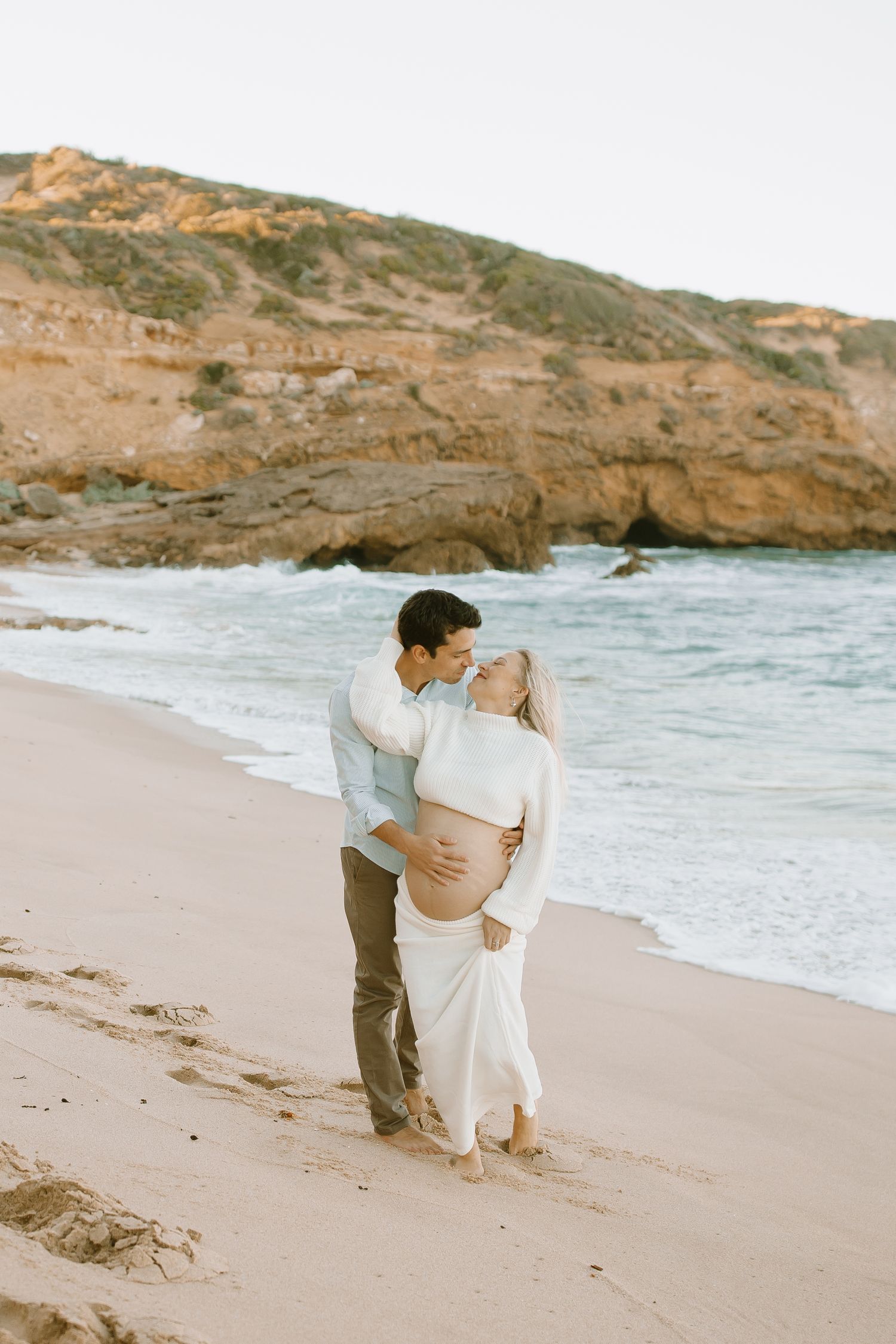A couple shares an intimate moment on a beach with rugged coastal cliffs in the background during golden hour.
