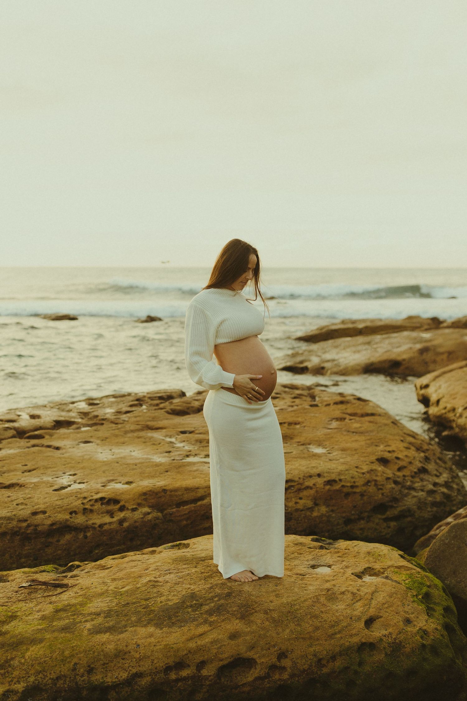 Expecting mother in white dress stands on rocky coastal outcrop at sunset while cradling her belly.