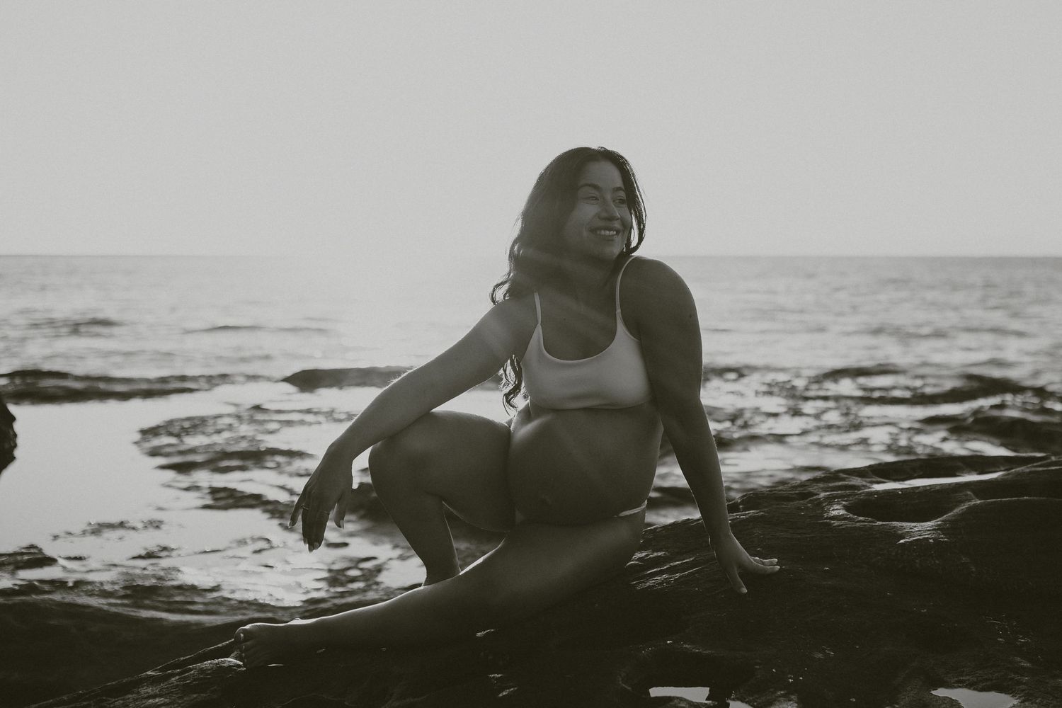 A person in a white top sits on dark rocks at the beach during sunset, creating a moody black and white scene.