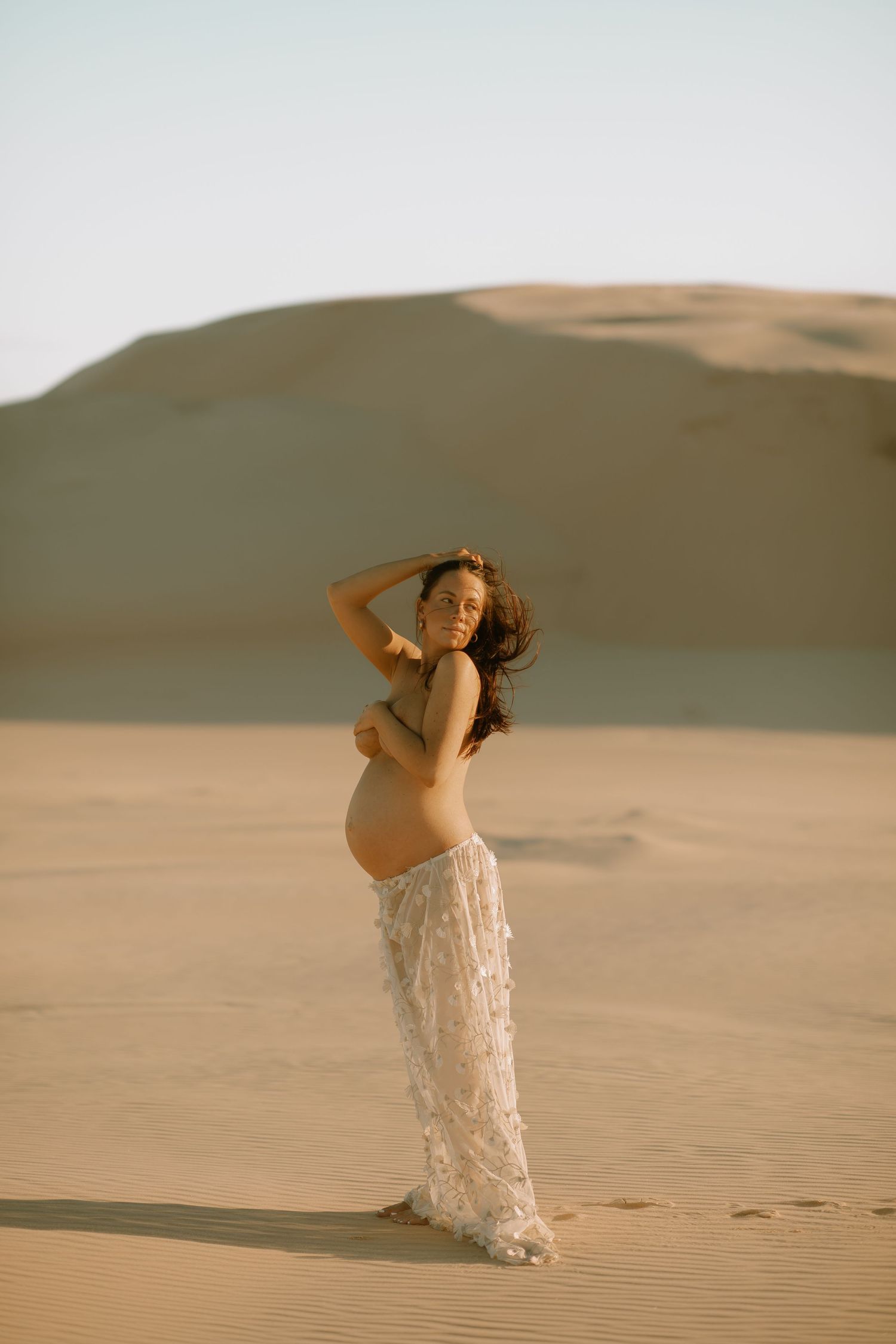 Ethereal maternity photoshoot in flowing white dress against sand dunes during golden hour.