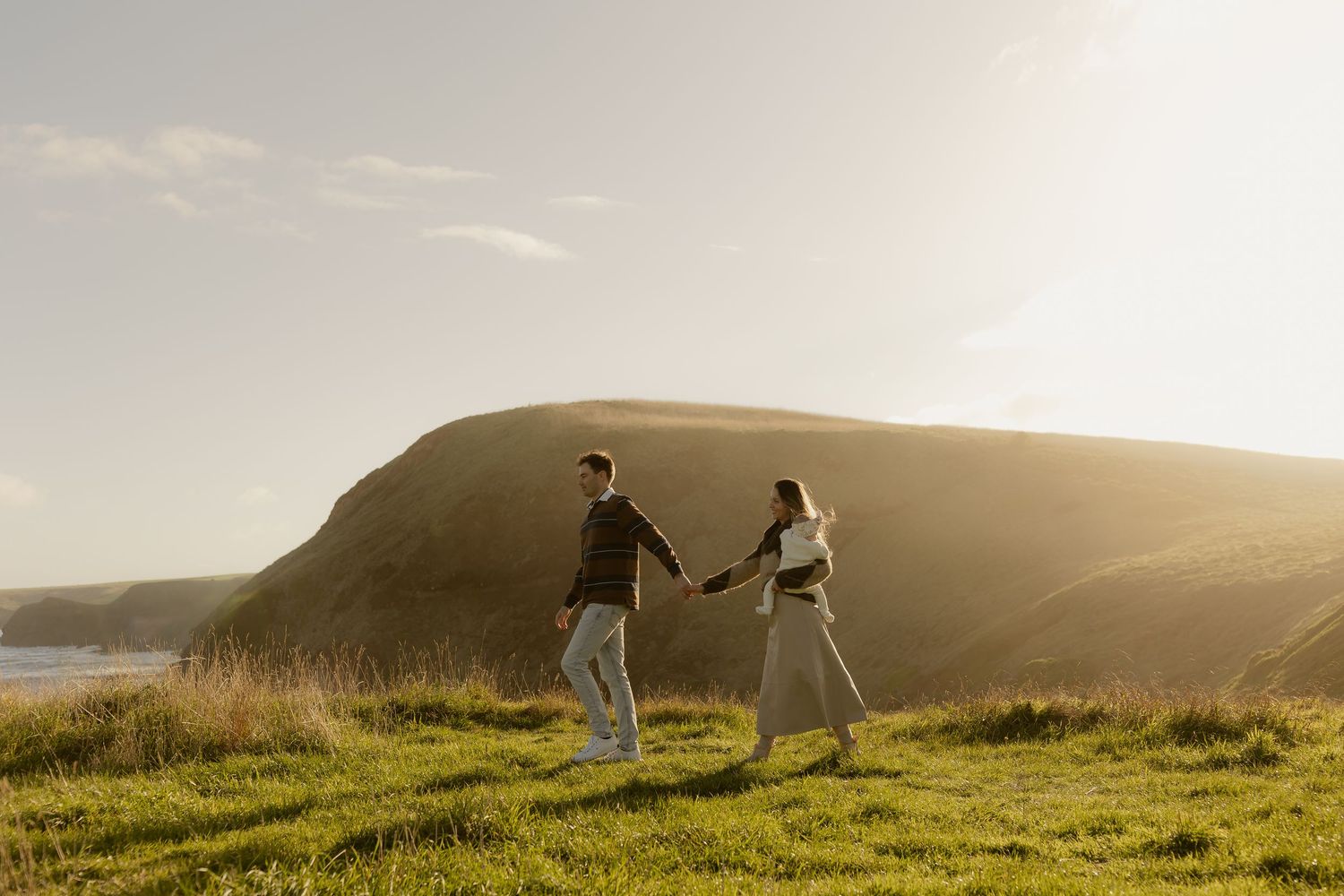 Couple holding hands walking on green coastal cliffs at golden hour sunset.