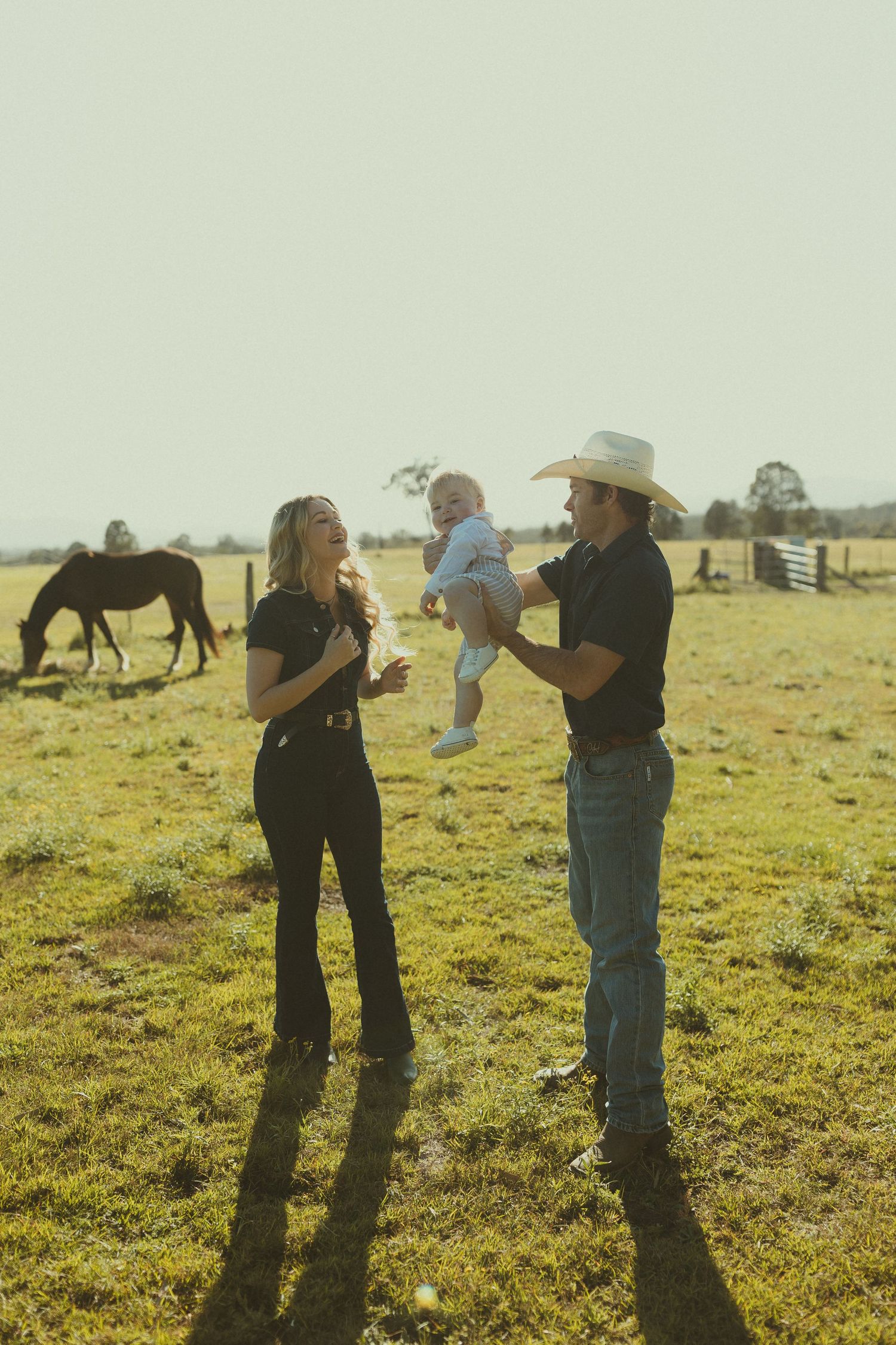 A family stands together in a sunny pasture with a horse grazing in the background on a warm afternoon.