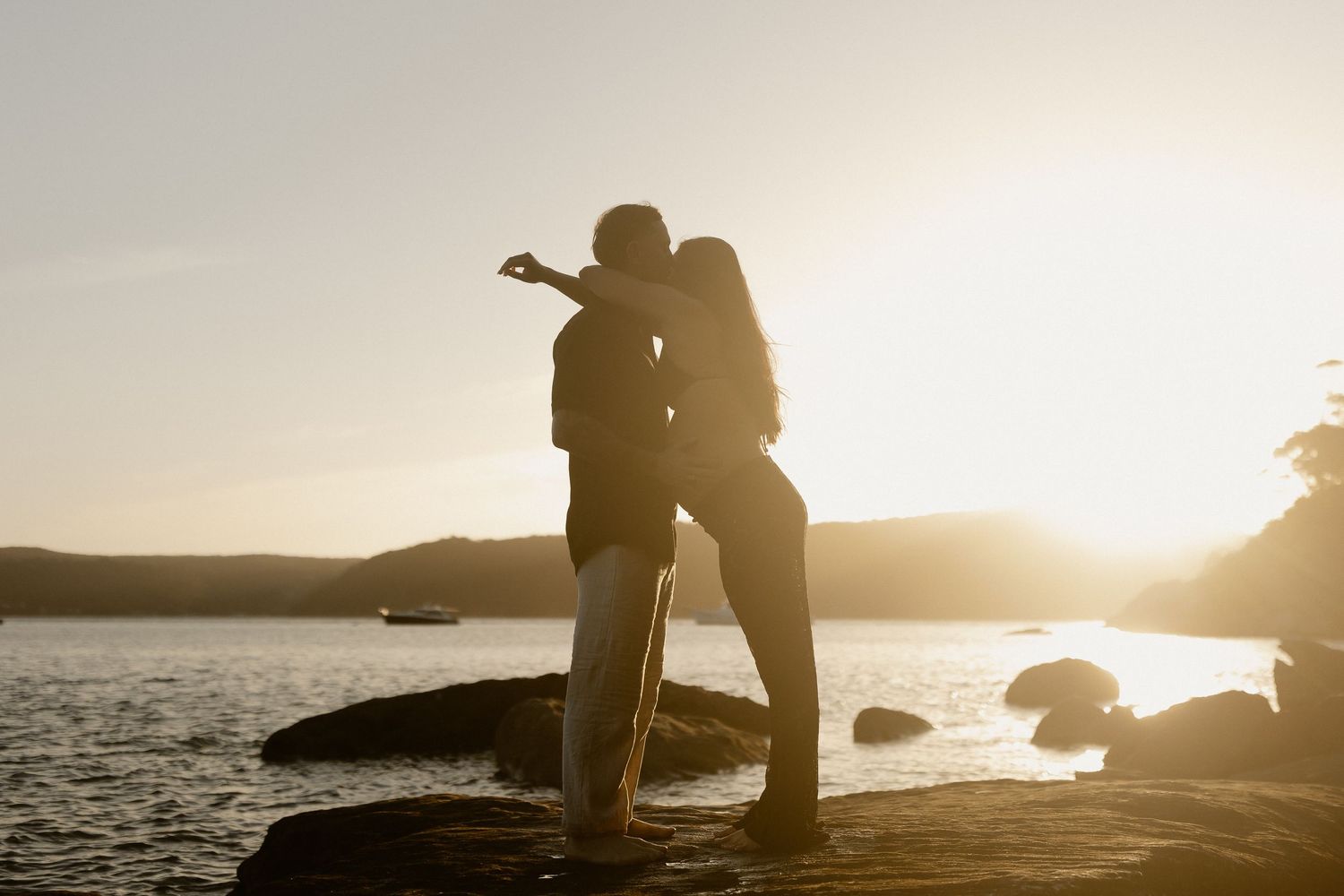 A silhouetted couple embraces on a rocky coastline during a golden sunset over the ocean.