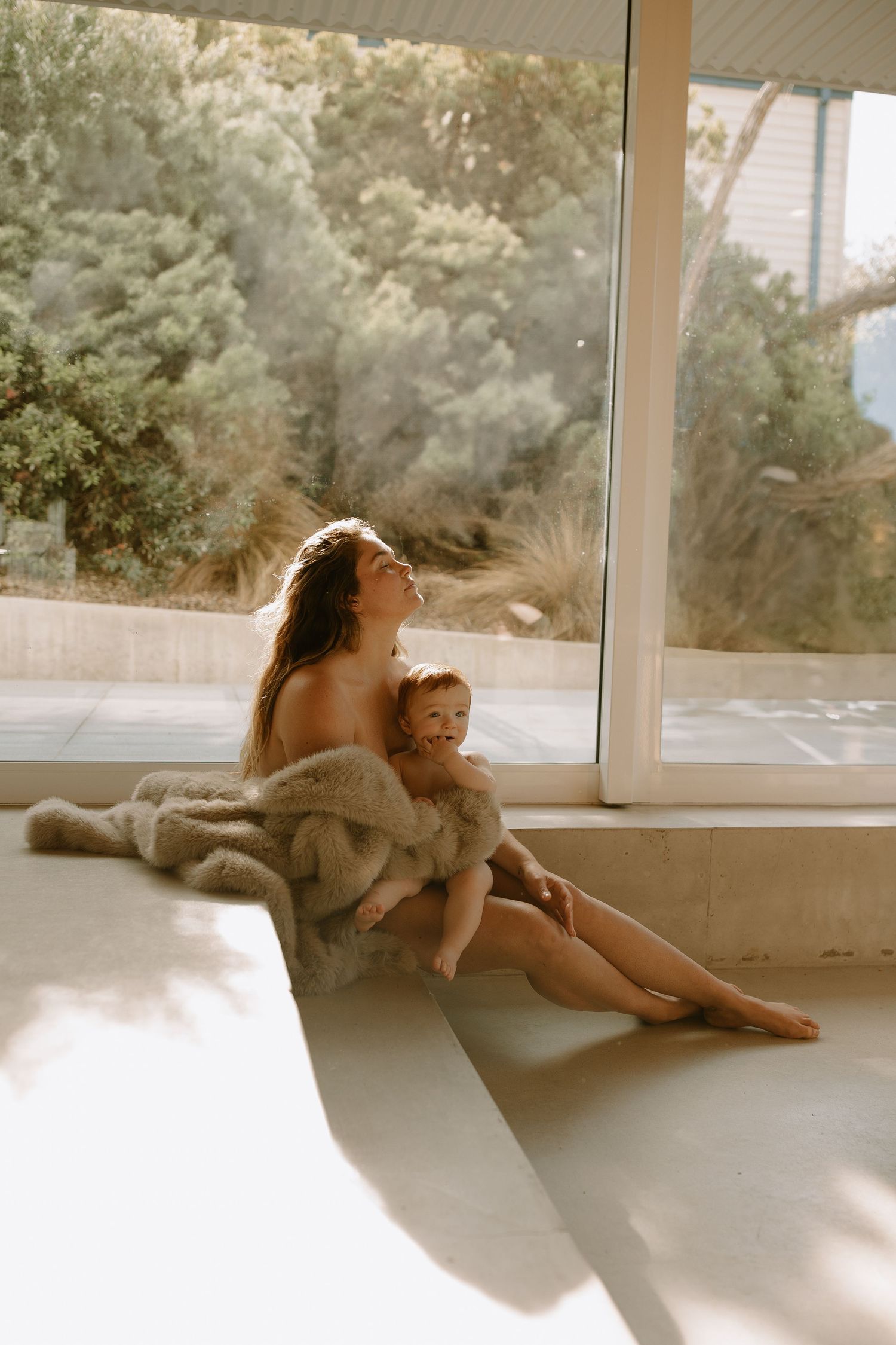 A tender moment is captured in warm sunlight streaming through a large window as two figures relax together on the floor.