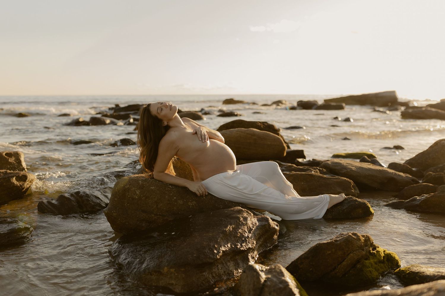 Artistic maternity photo of a person reclining on coastal rocks at golden hour.
