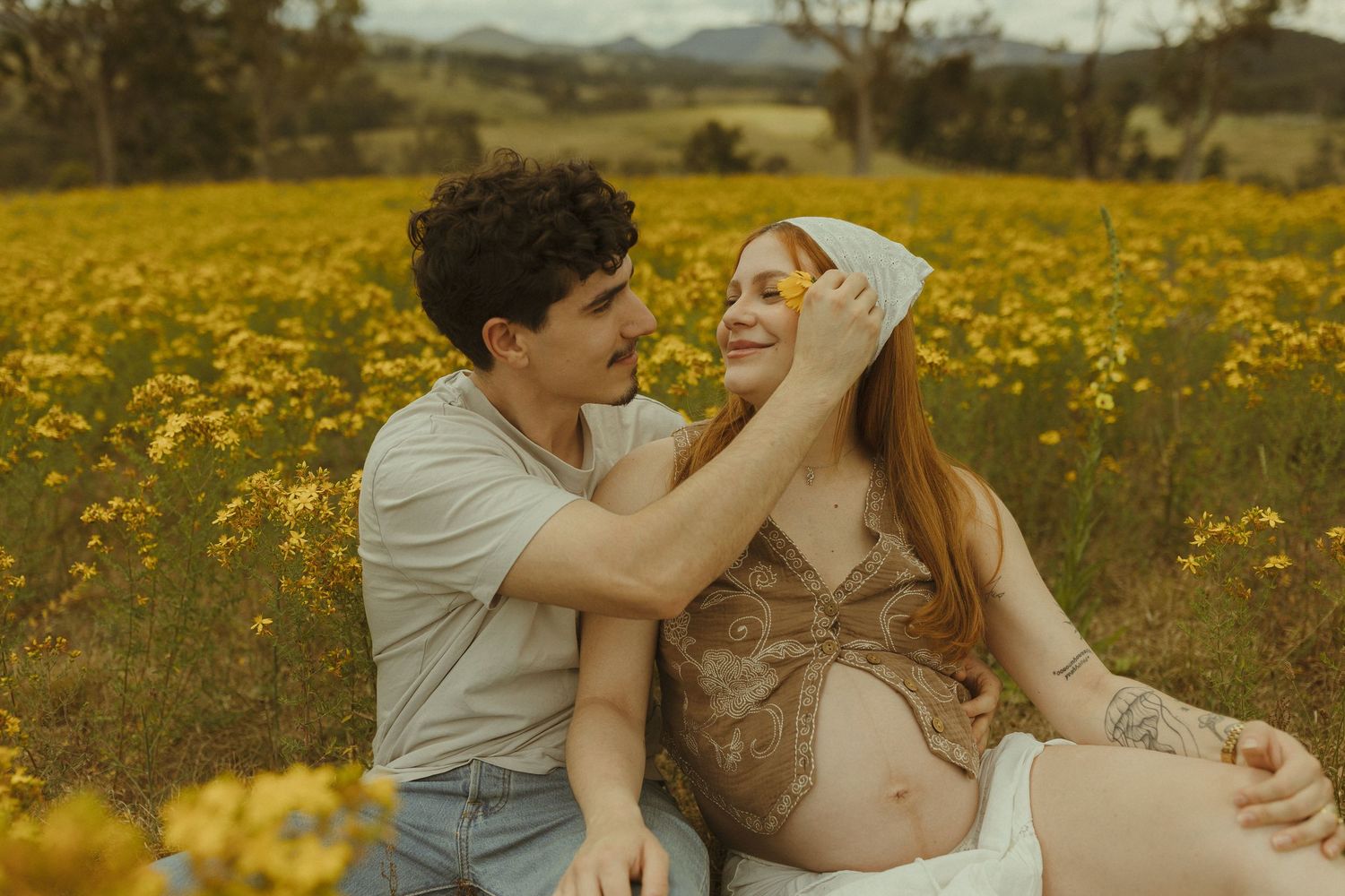 A couple embraces in a golden field of wildflowers during sunset with mountains in the background.