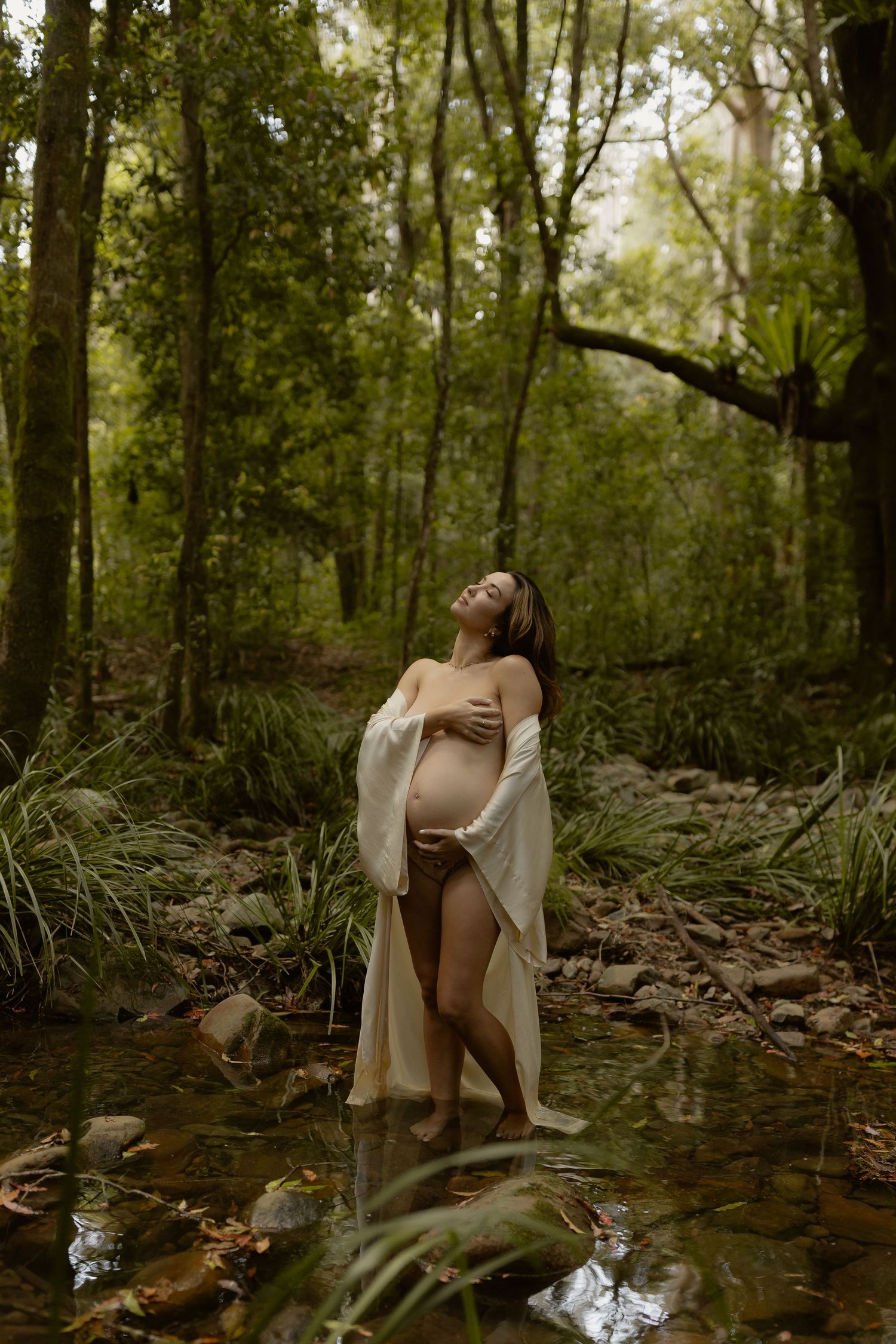 Pregnant woman in sheer wrap stands in forest stream, head tilted back.