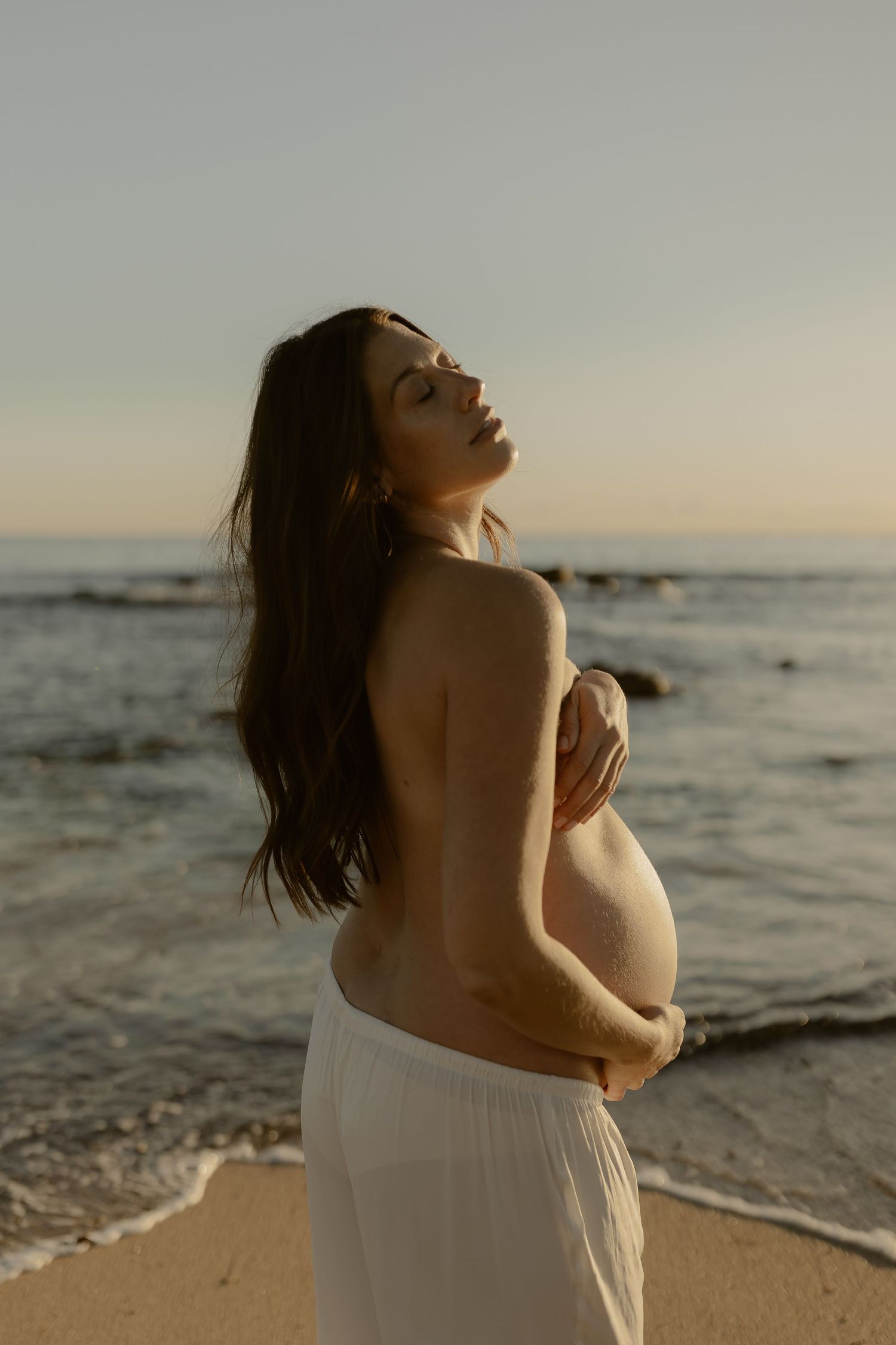 A pregnant woman stands topless on a beach at sunset, eyes closed, embracing her baby bump in white skirt.