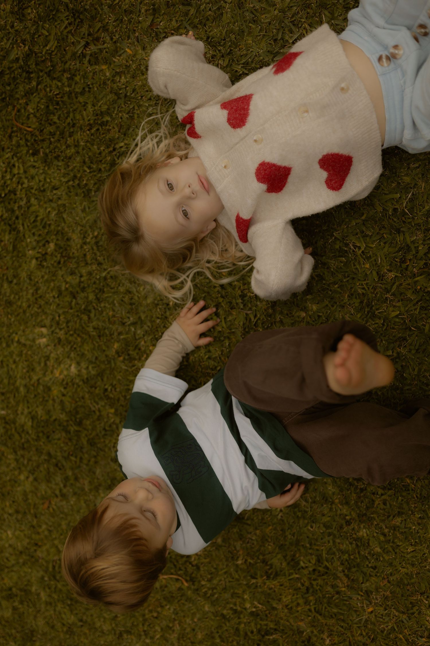 Two young children lie on green grass from above, a girl in a heart sweater and boy in a striped shirt.