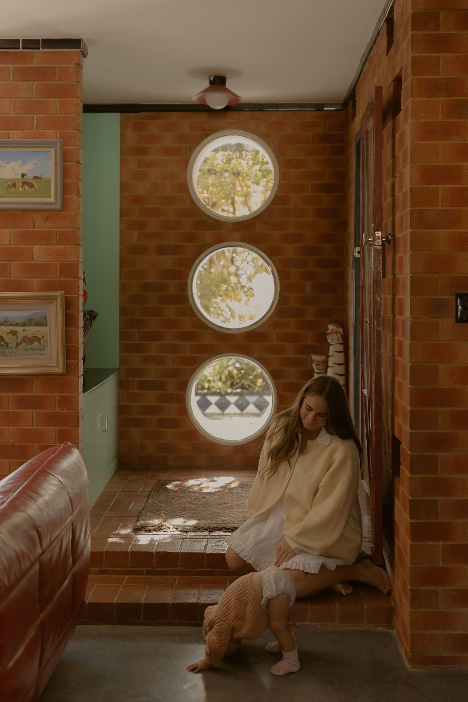 Woman sitting by brick wall with circular porthole windows casting light patterns on terracotta tile floor.