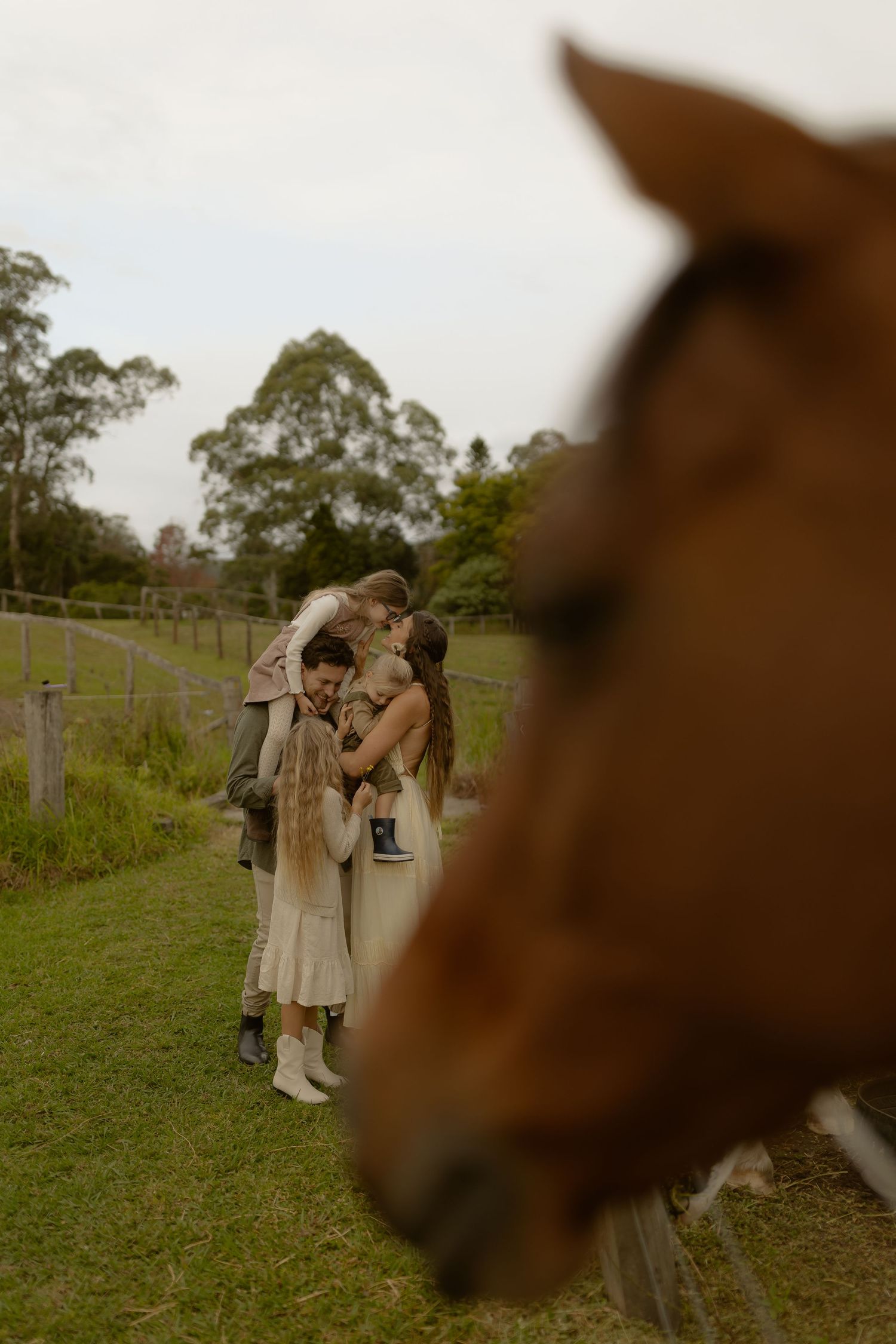 Mother and two children sharing a tender moment on a farm with a brown horse in the foreground.