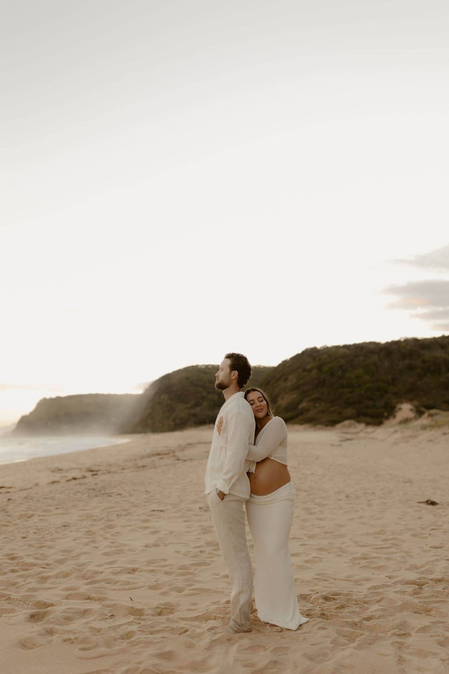 A couple in white outfits shares an intimate moment on a sandy beach at sunset.