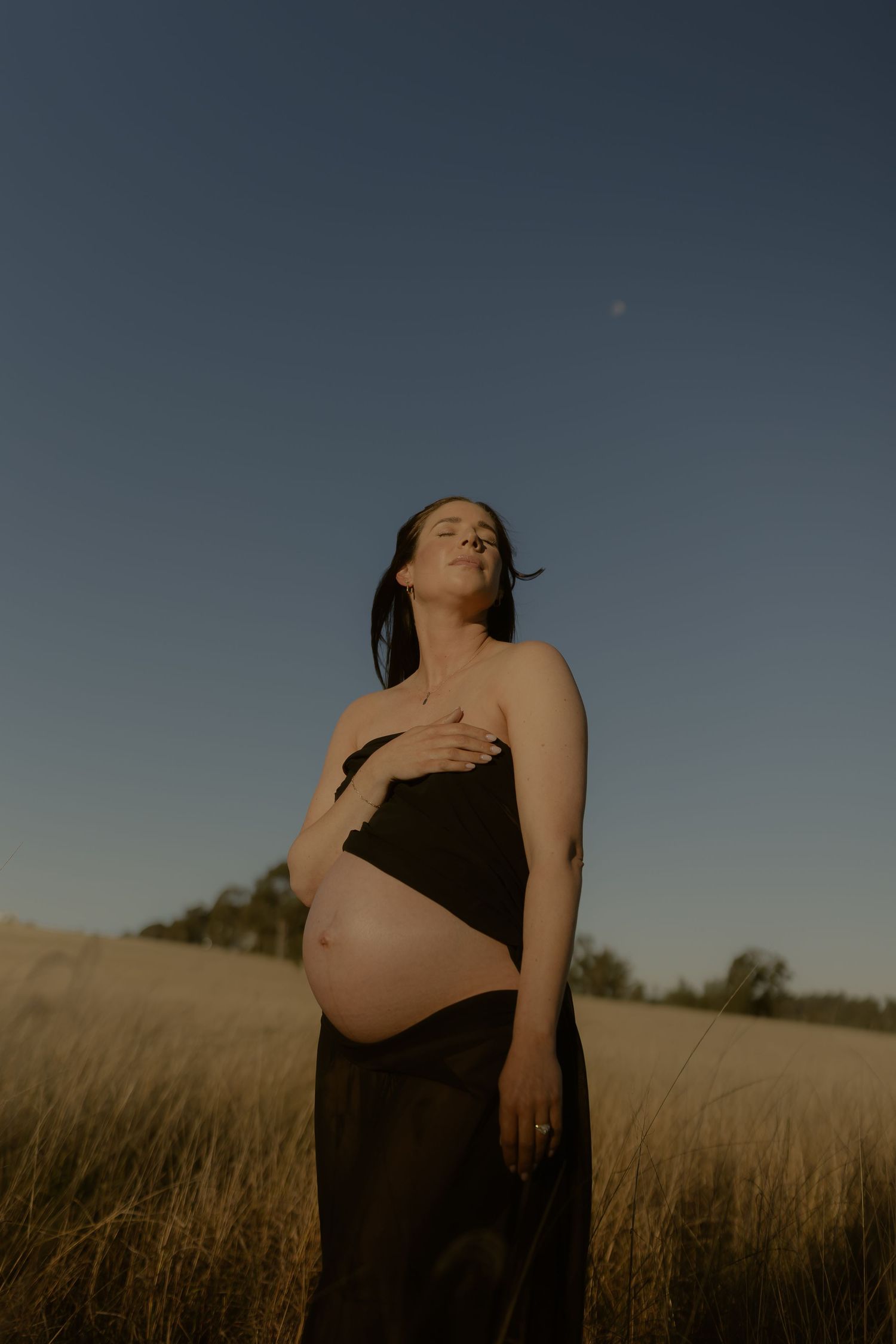 Pregnant woman in black wrap standing in golden field at dusk under clear blue sky.