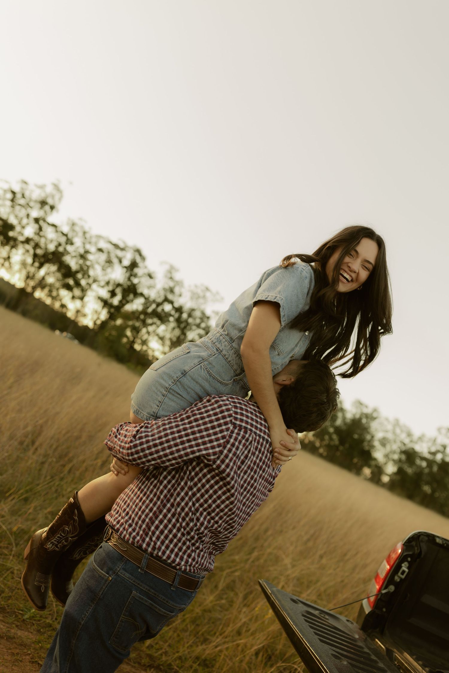A laughing woman is lifted over a mans shoulder in a golden field near a pickup truck at sunset.