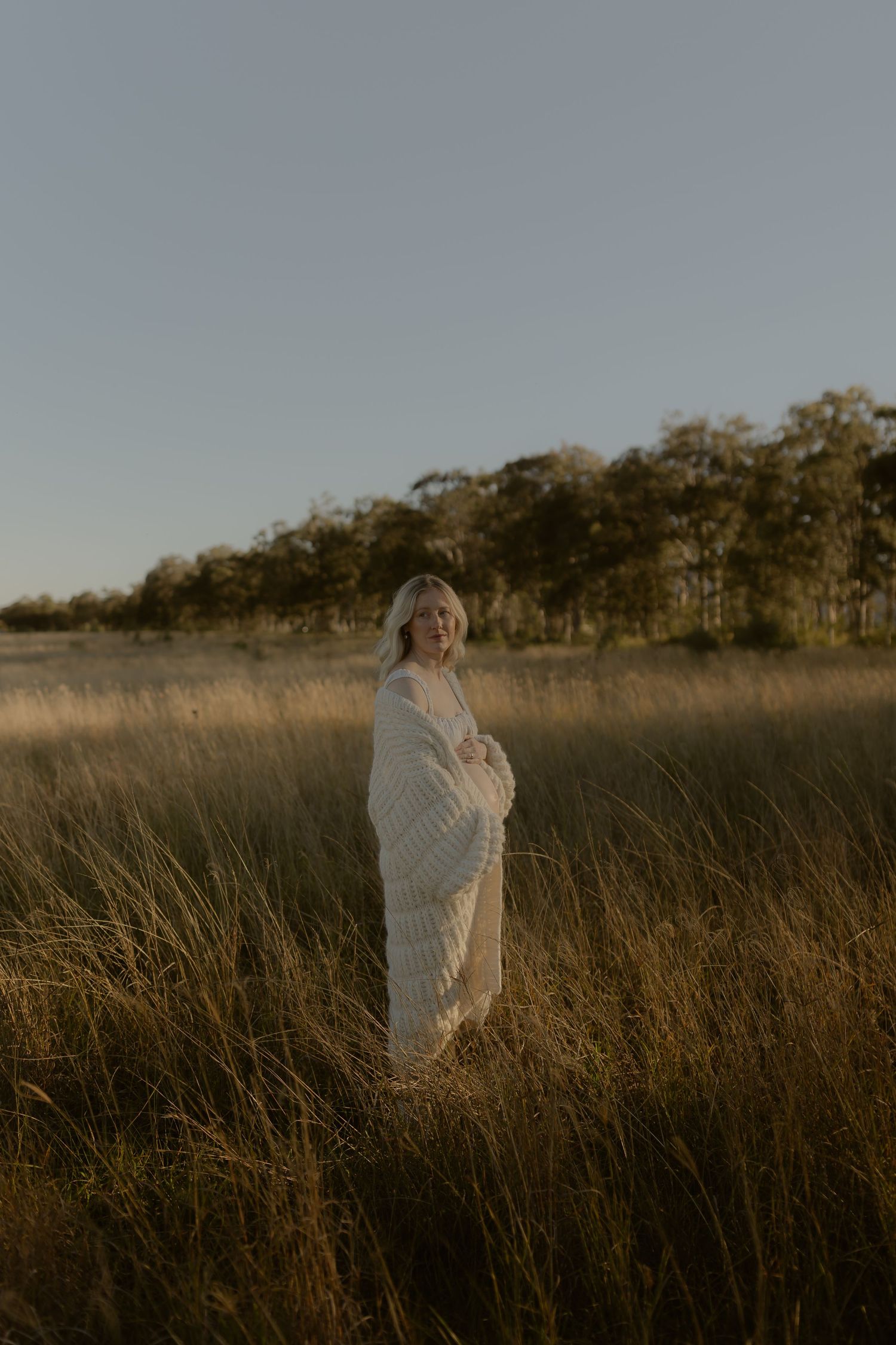 Woman wrapped in white blanket standing in golden grass field at sunset with trees in background.