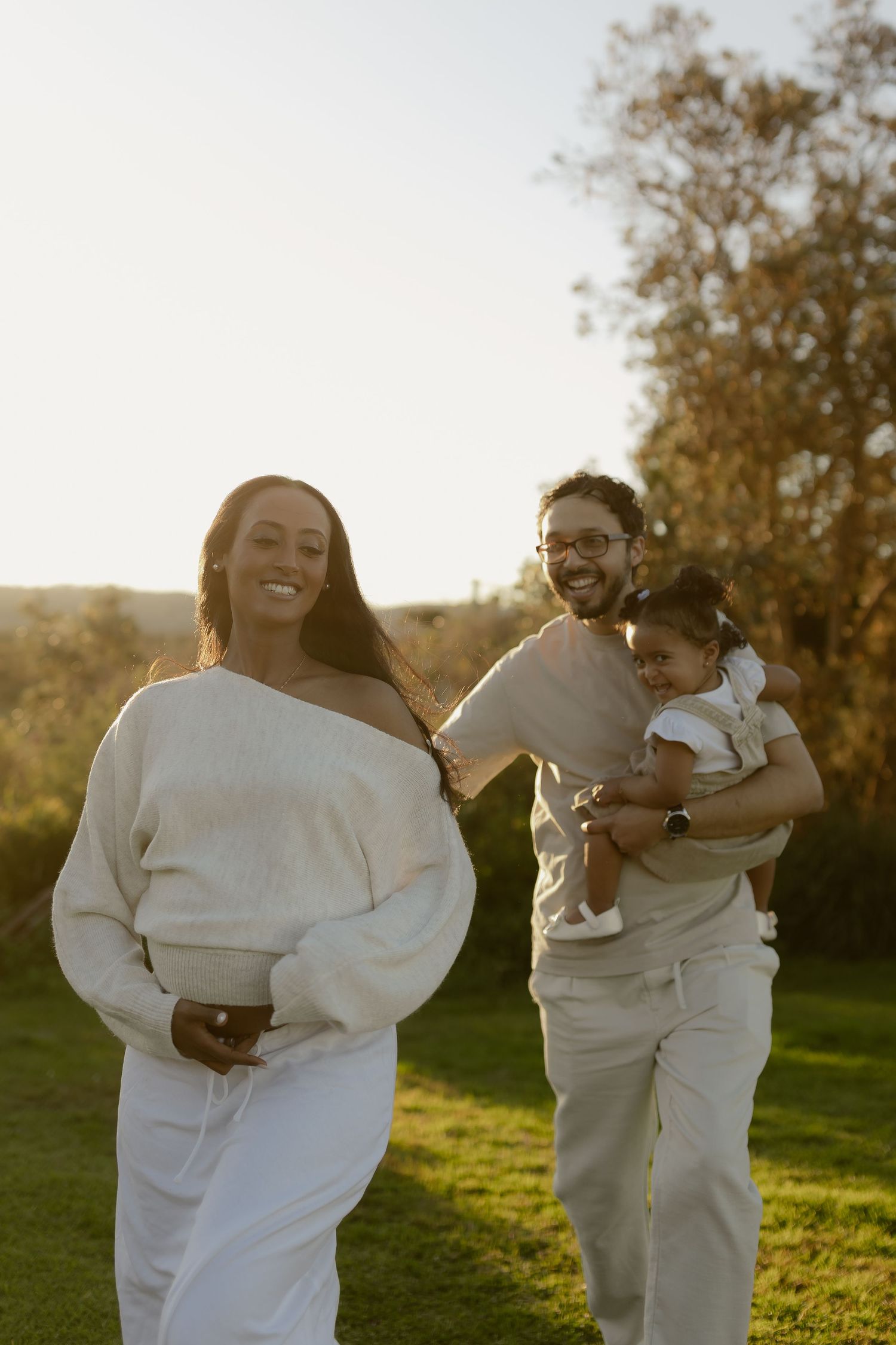 Happy family walking outdoors at golden hour, mother in white sweater, father holding baby girl.