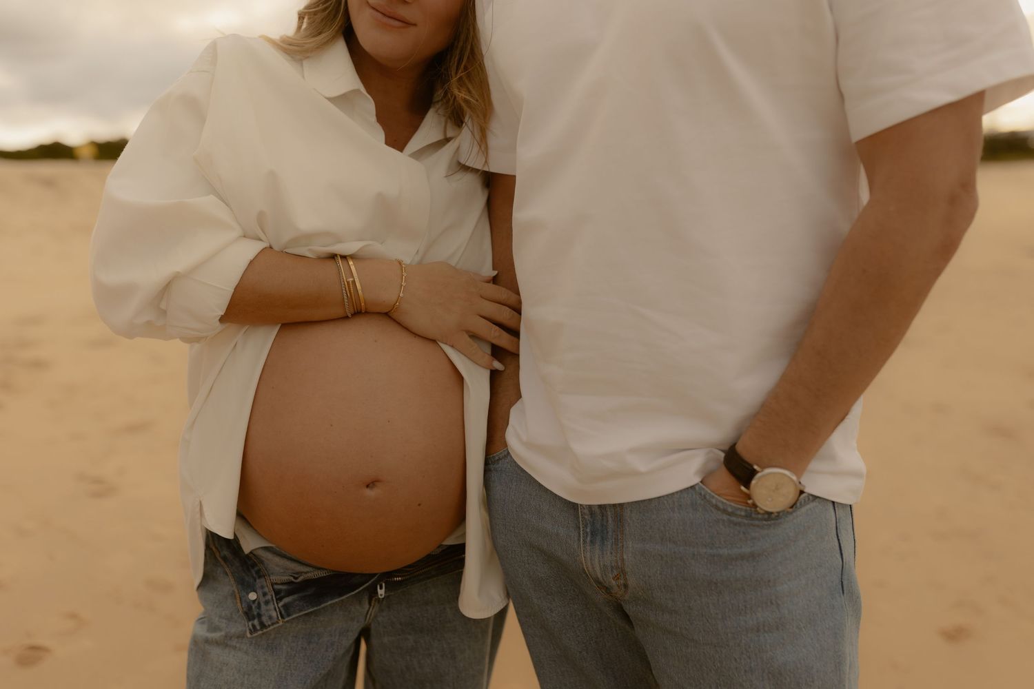 Pregnant couple standing on sandy beach, woman in white shirt with exposed baby bump, man in white tee and jeans beside her.