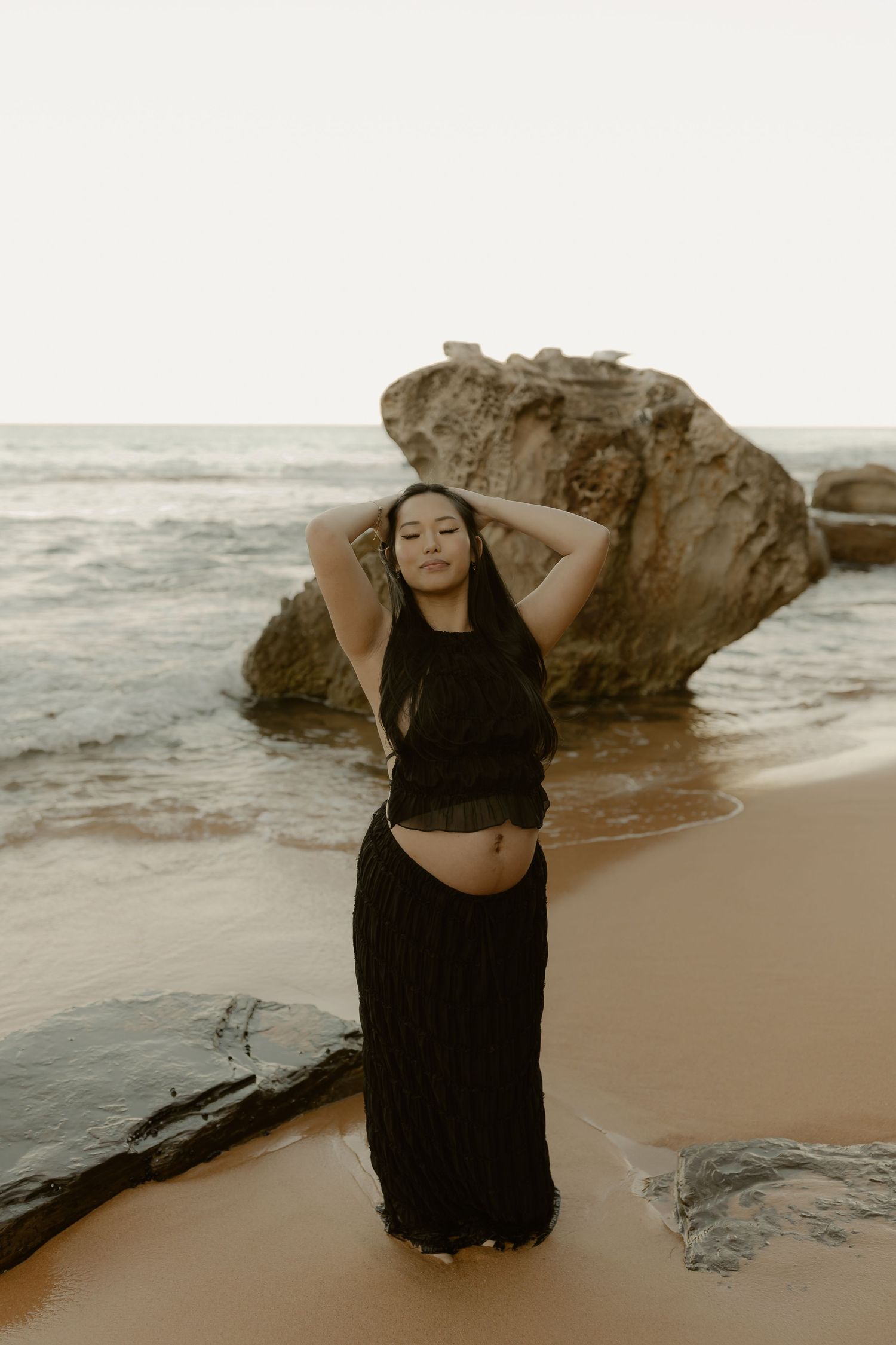 A woman in a black crop top and maxi skirt poses on a sandy beach with rocky formations and ocean waves behind her.