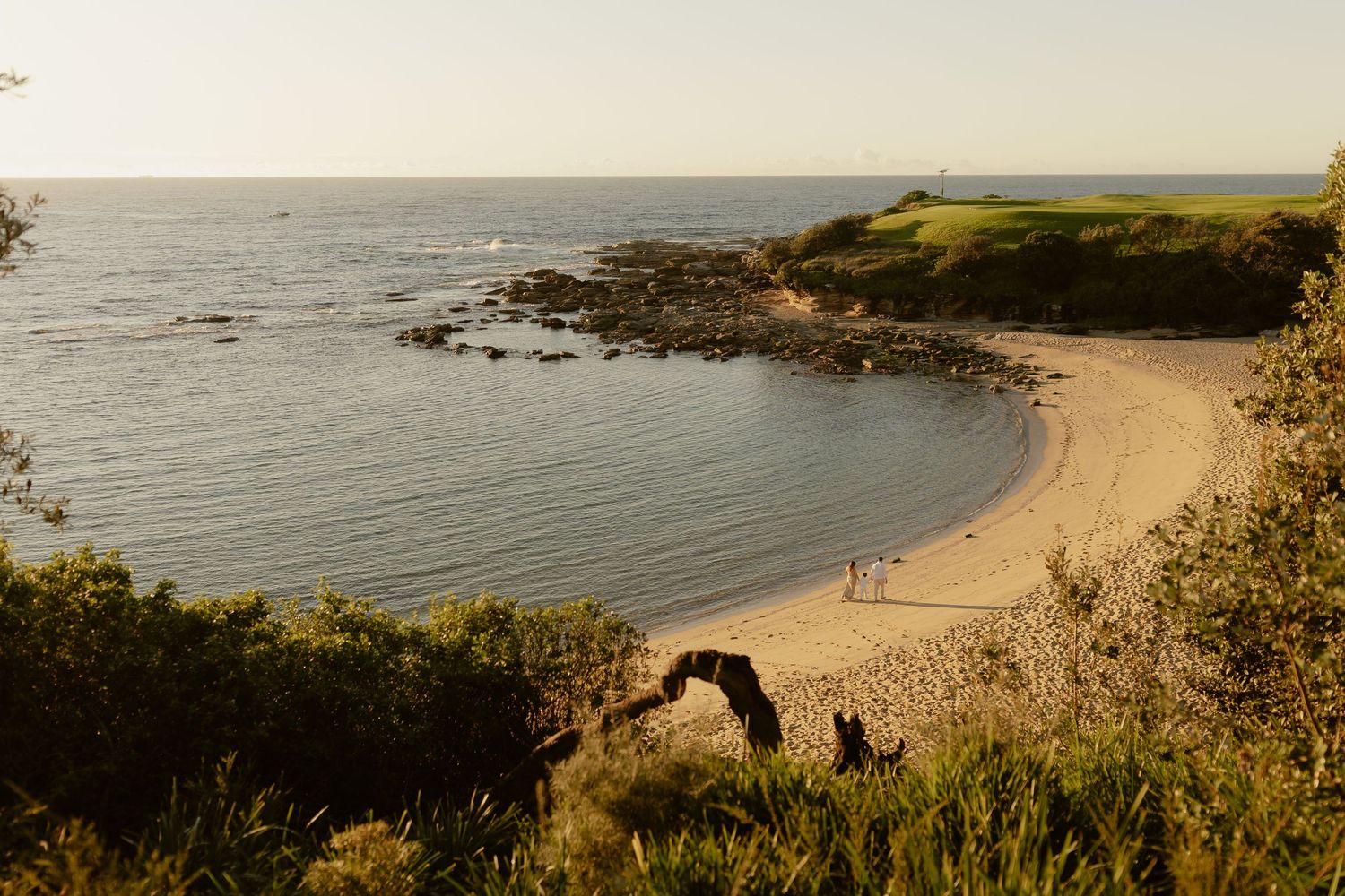 Aerial view of a secluded sandy beach cove with calm blue waters, rocky shores, and lush green coastal vegetation.