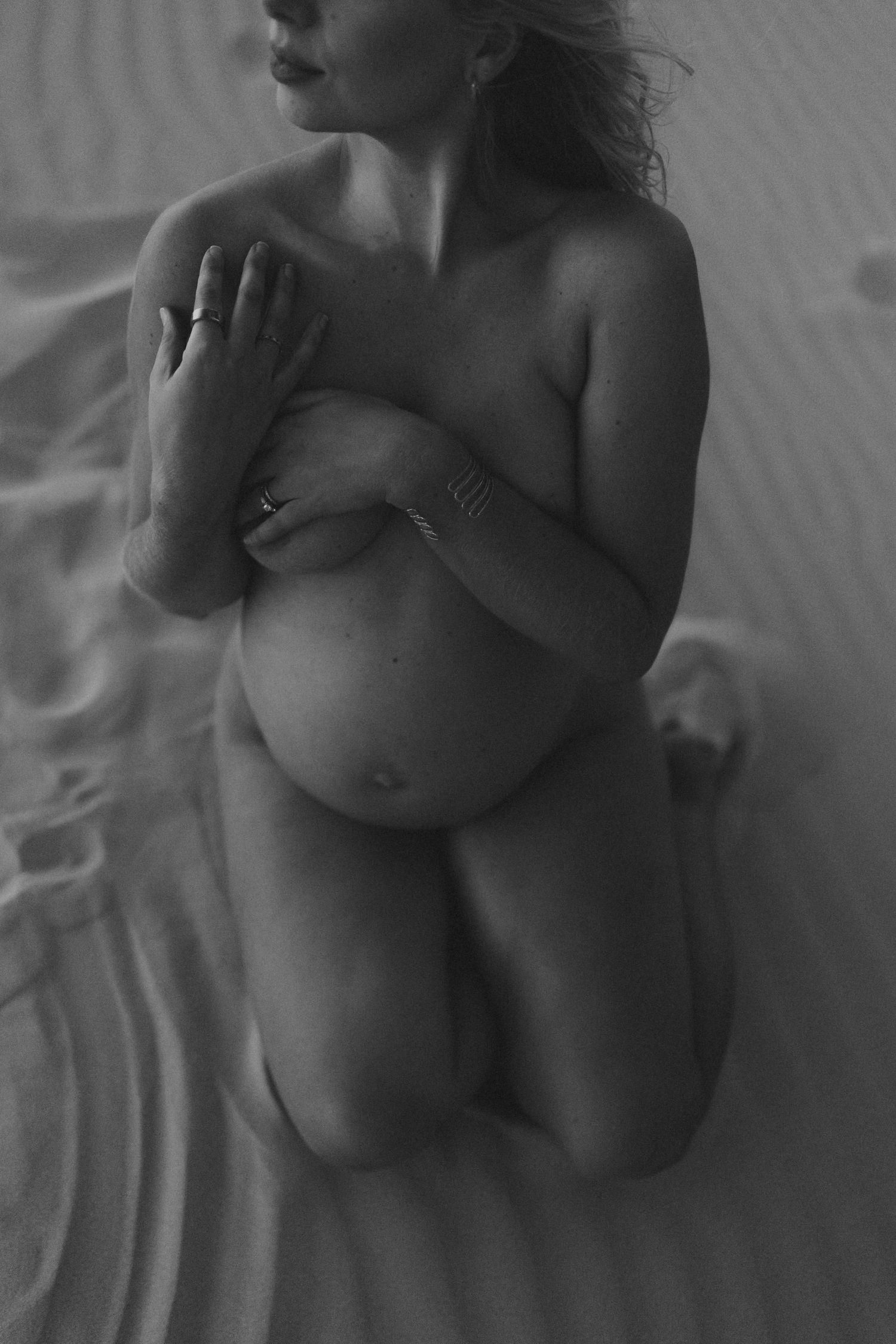 Black and white artistic photo of a pregnant woman kneeling on sand, arms crossed over chest.