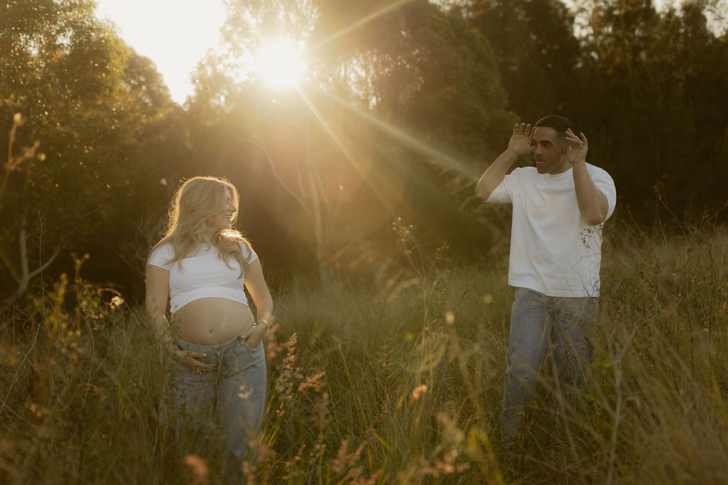 Expecting couple standing apart in a sun-drenched meadow at golden hour with lens flare.