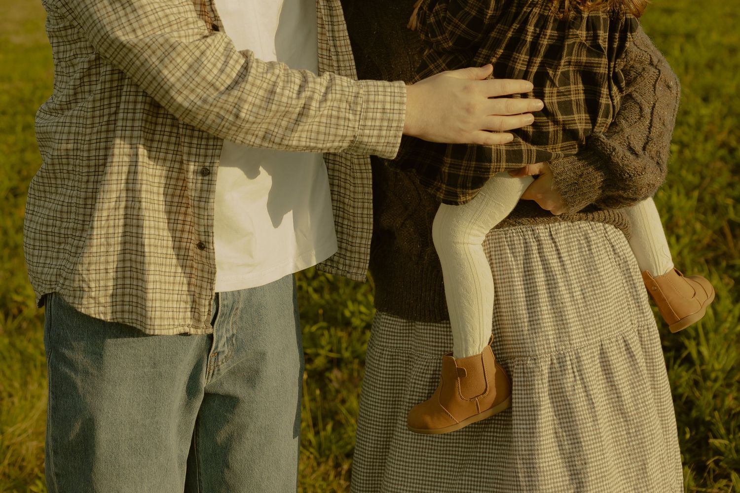 Family holding a young child outdoors in a sunlit green field, wearing casual plaid and neutral tones.