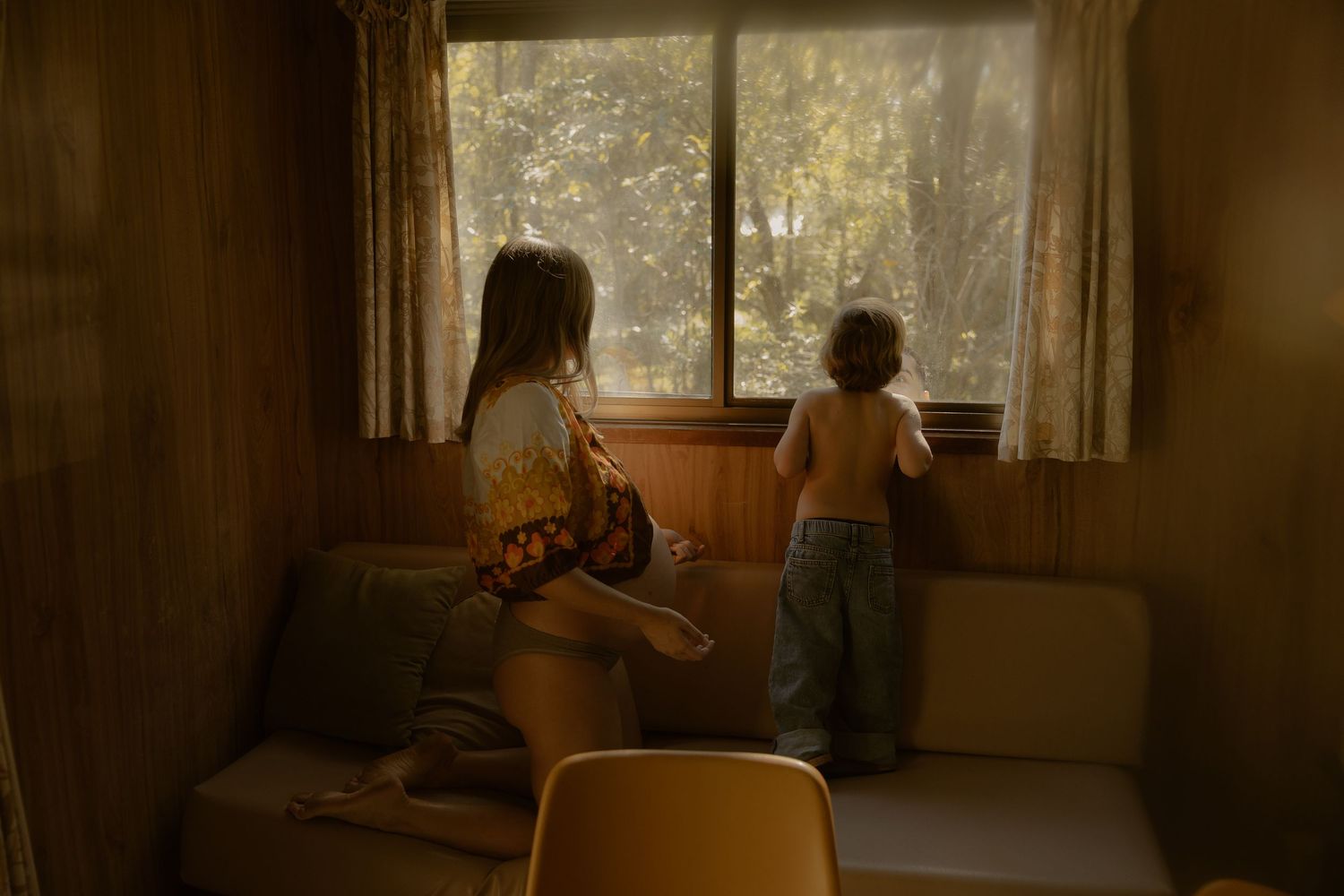 Two young children look out a window in a warmly lit, wood-paneled room, one standing and one sitting on a couch.