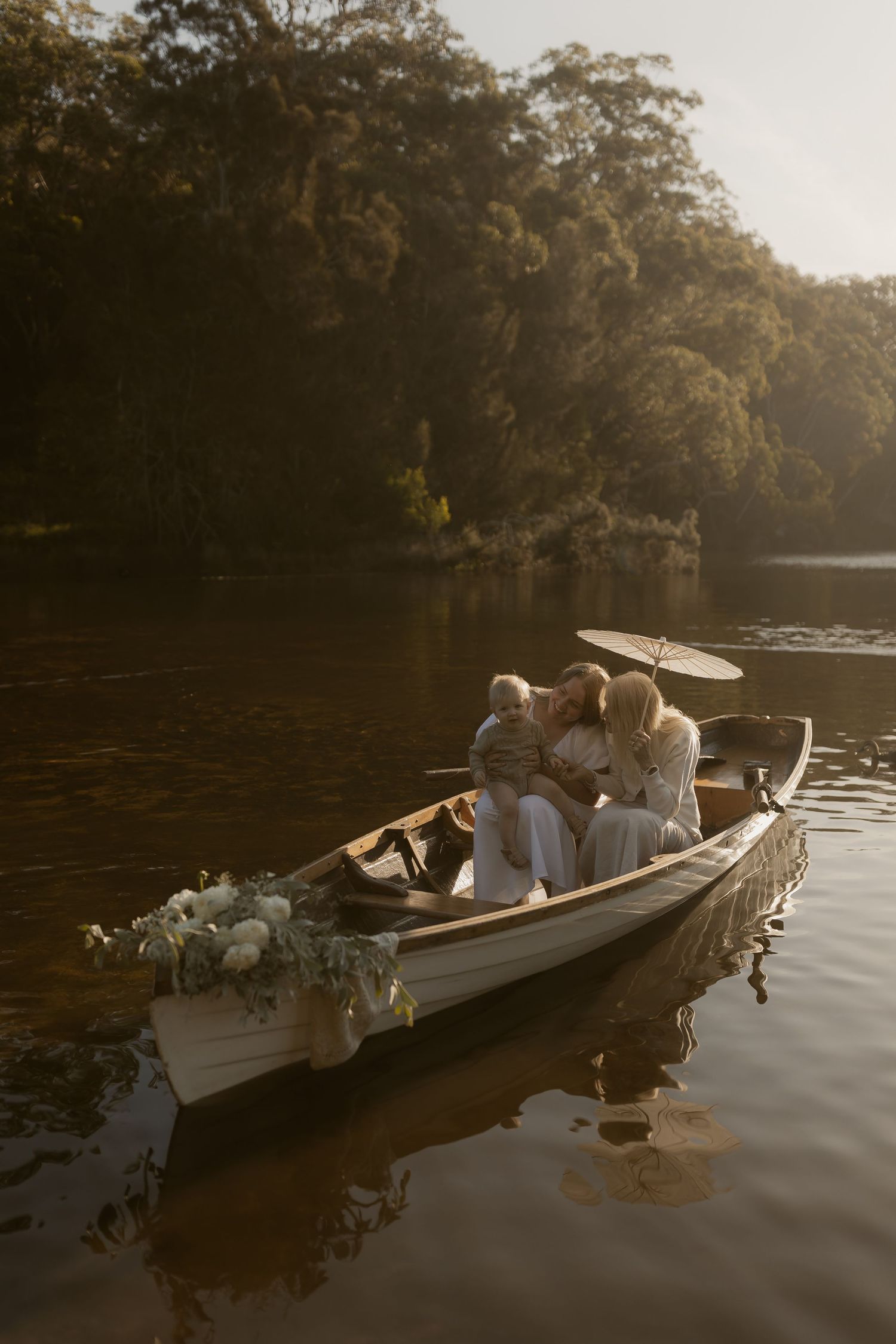 Family with child sitting in a decorated white rowboat on a calm river surrounded by lush green trees at golden hour.