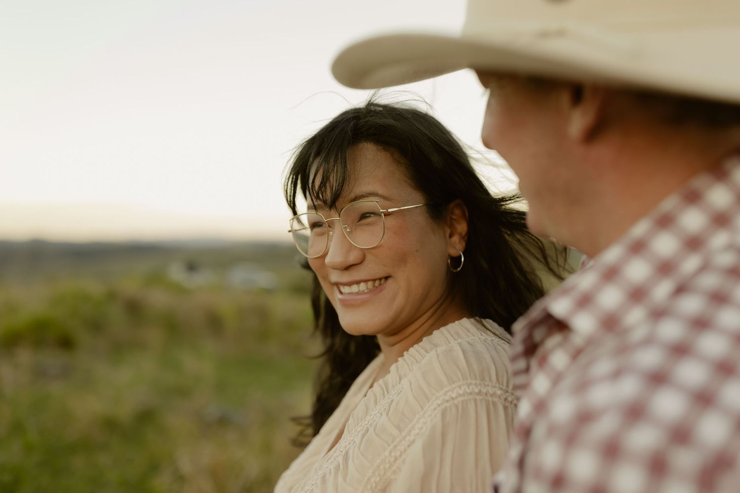 A smiling woman with glasses and dark hair stands outdoors near a man wearing a cowboy hat at sunset.