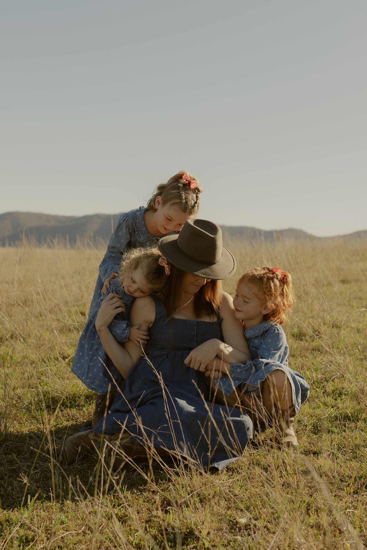 A mother in a cowboy hat crouches in a golden field, tenderly embracing her three young daughters outdoors.