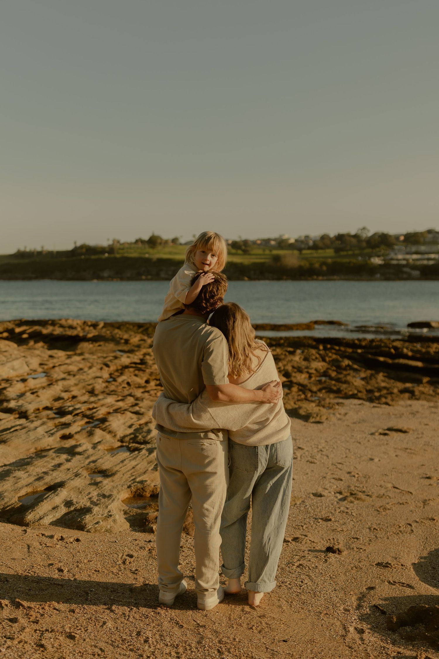 Two women embracing on a sandy beach at golden hour, gazing at the ocean with rocky shores and greenery in the background.