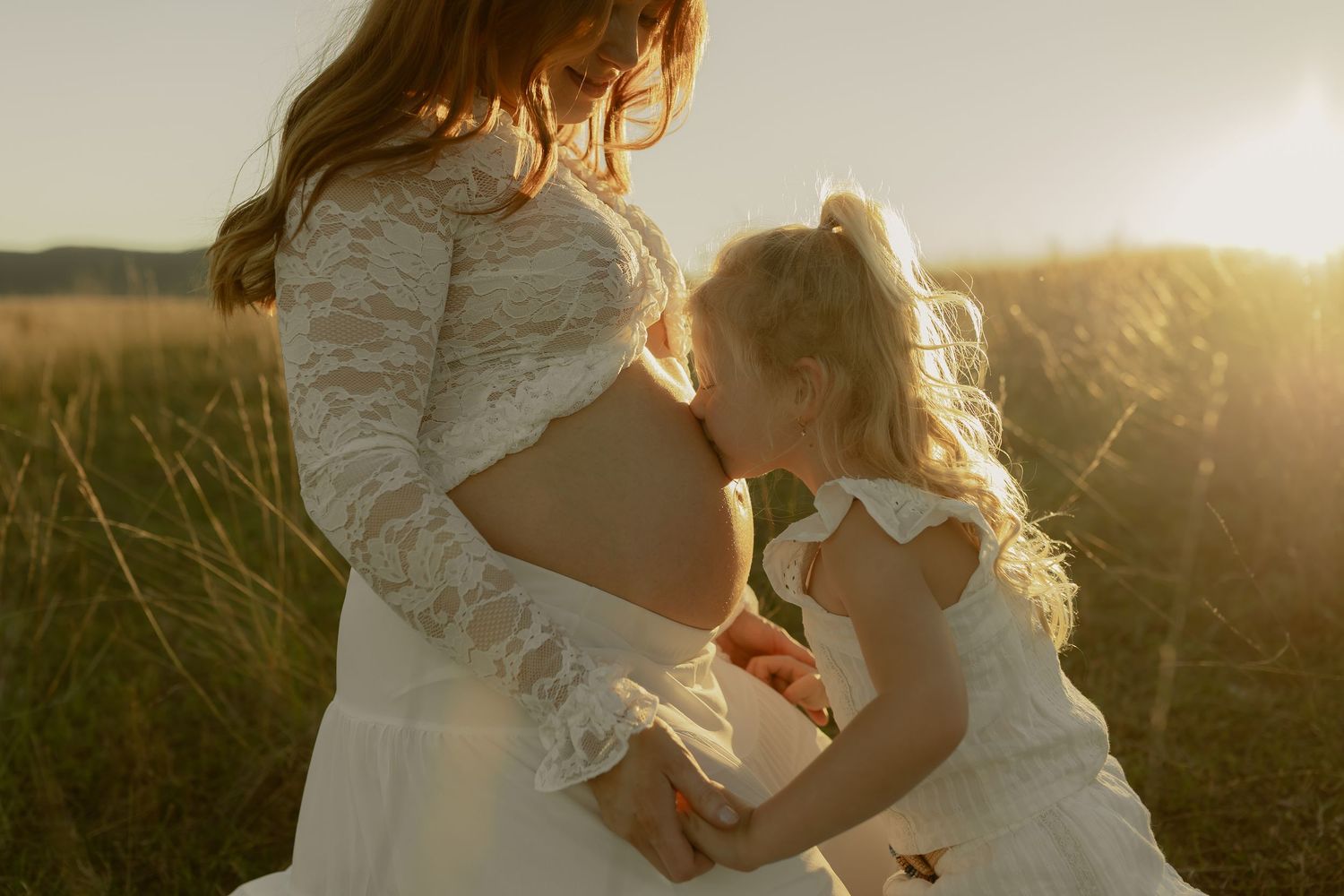A young girl in a white dress kisses a pregnant woman's belly in a golden sunset field.