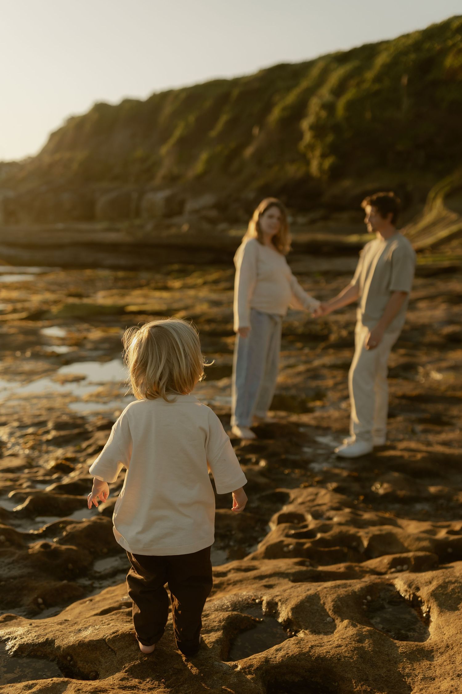 A young child walks toward their parents holding hands on rocky coastal cliffs during golden hour sunset.