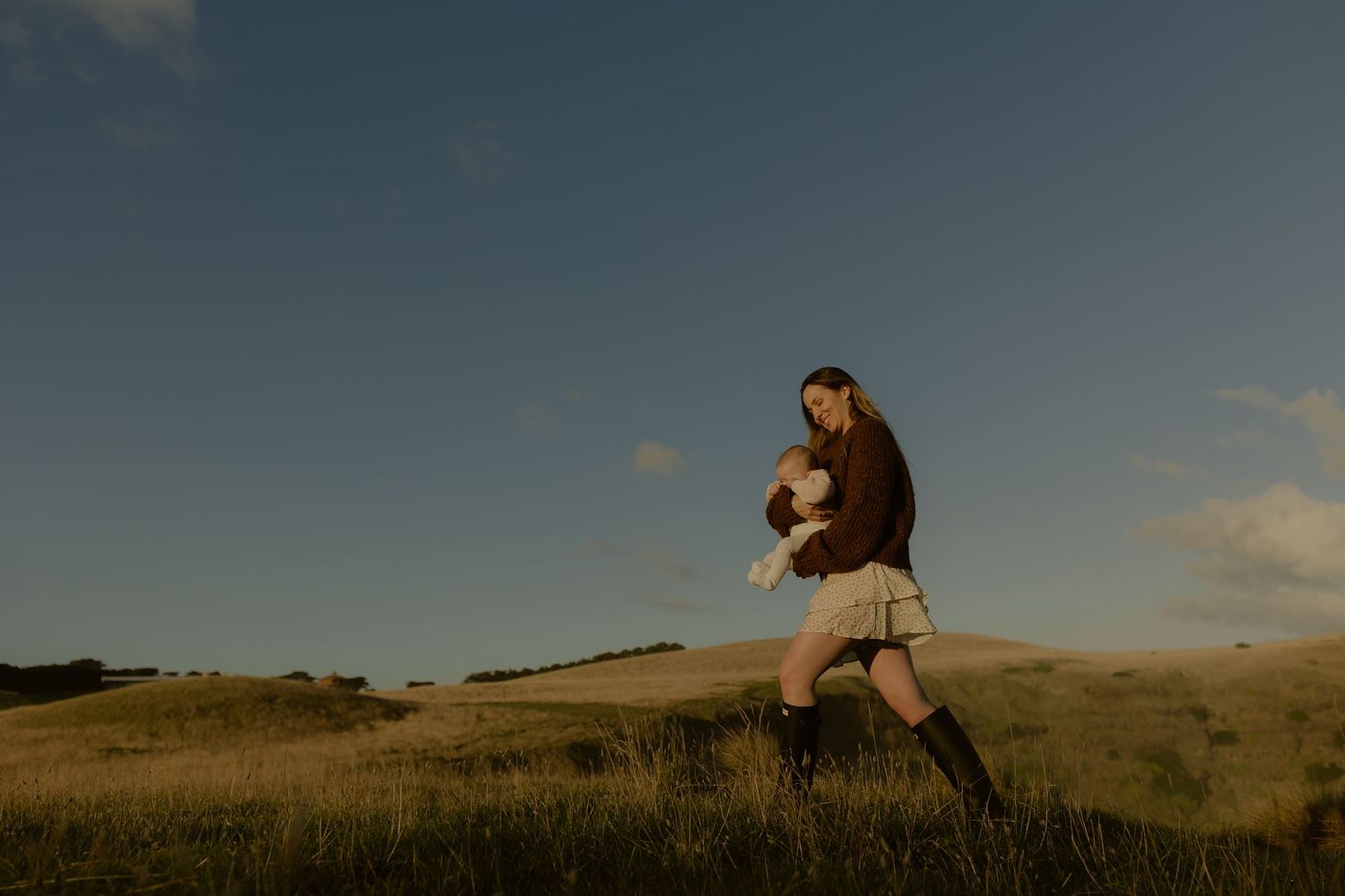 A person walks through a golden meadow under a blue sky, carrying a small child in warm afternoon light.
