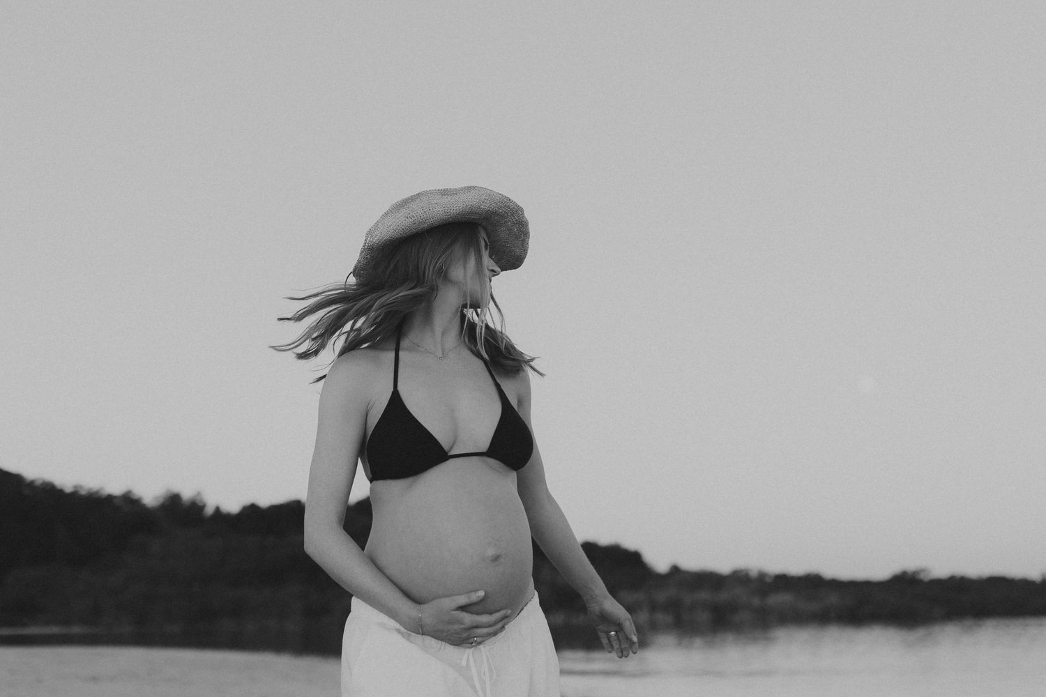 Pregnant woman in black bikini top and white skirt wearing a wide-brim hat stands near water in black and white photo.
