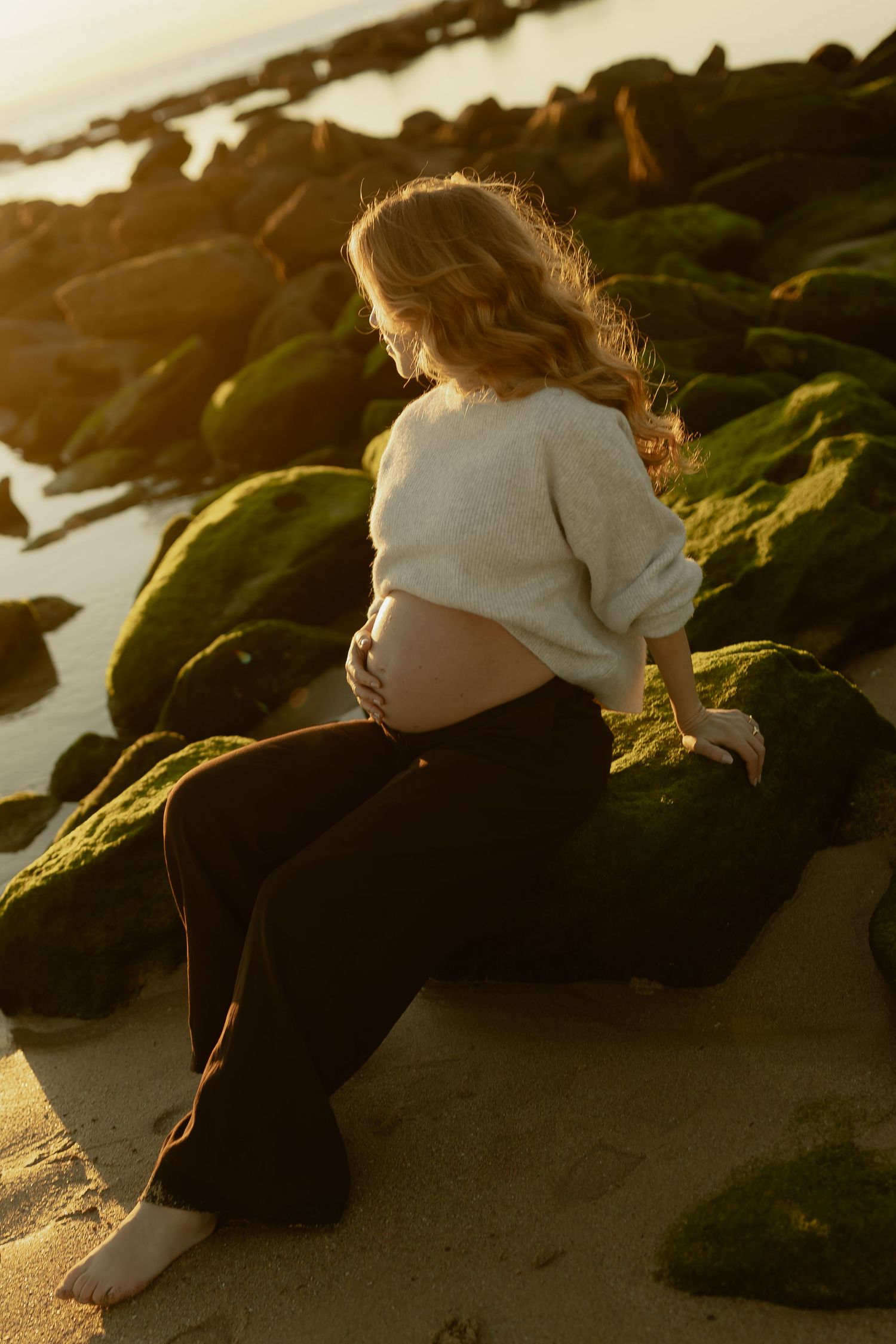 Pregnant woman in cream sweater sits on moss-covered rocks at beach during warm golden sunset.