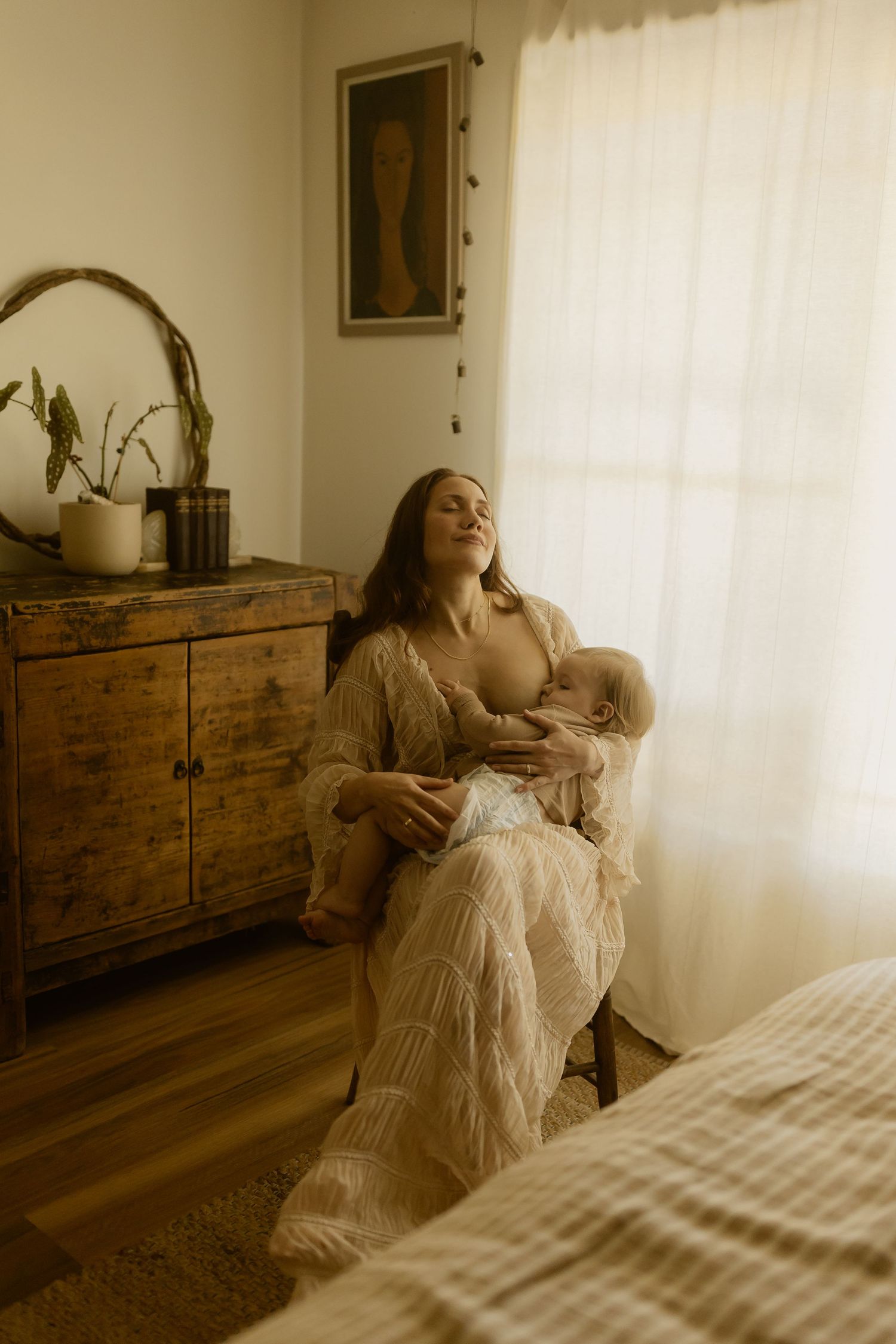 A tender nursing moment in a sunlit room with vintage wooden furniture and flowing white curtains.