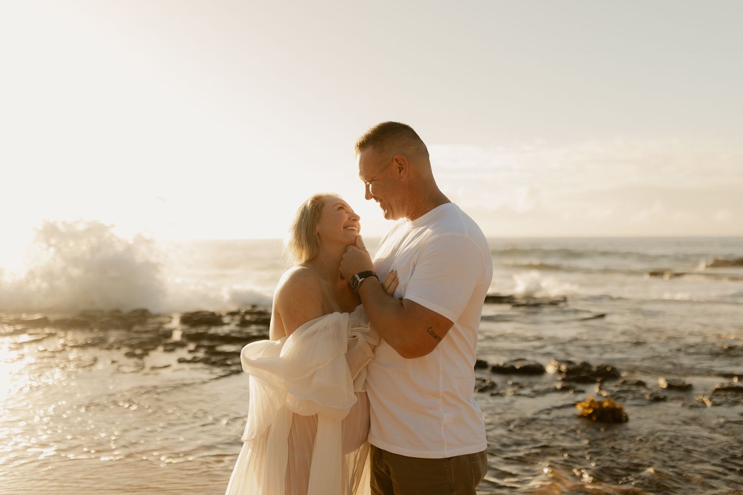 Couple embracing on a rocky beach at golden sunset with ocean waves crashing behind them.
