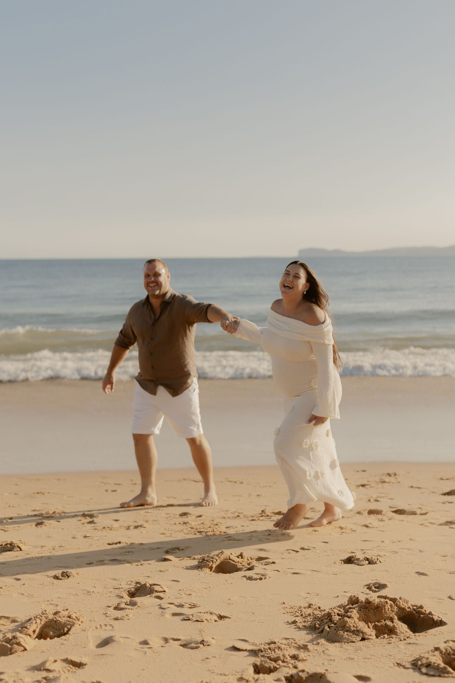 Playful couple running and dancing together on a sandy beach at sunset with ocean waves in the background.