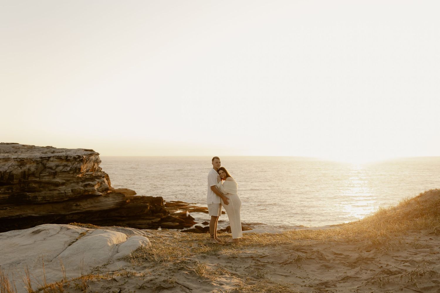 Scenic beach engagement photoshoot with rocky cliffs and golden sunset light.