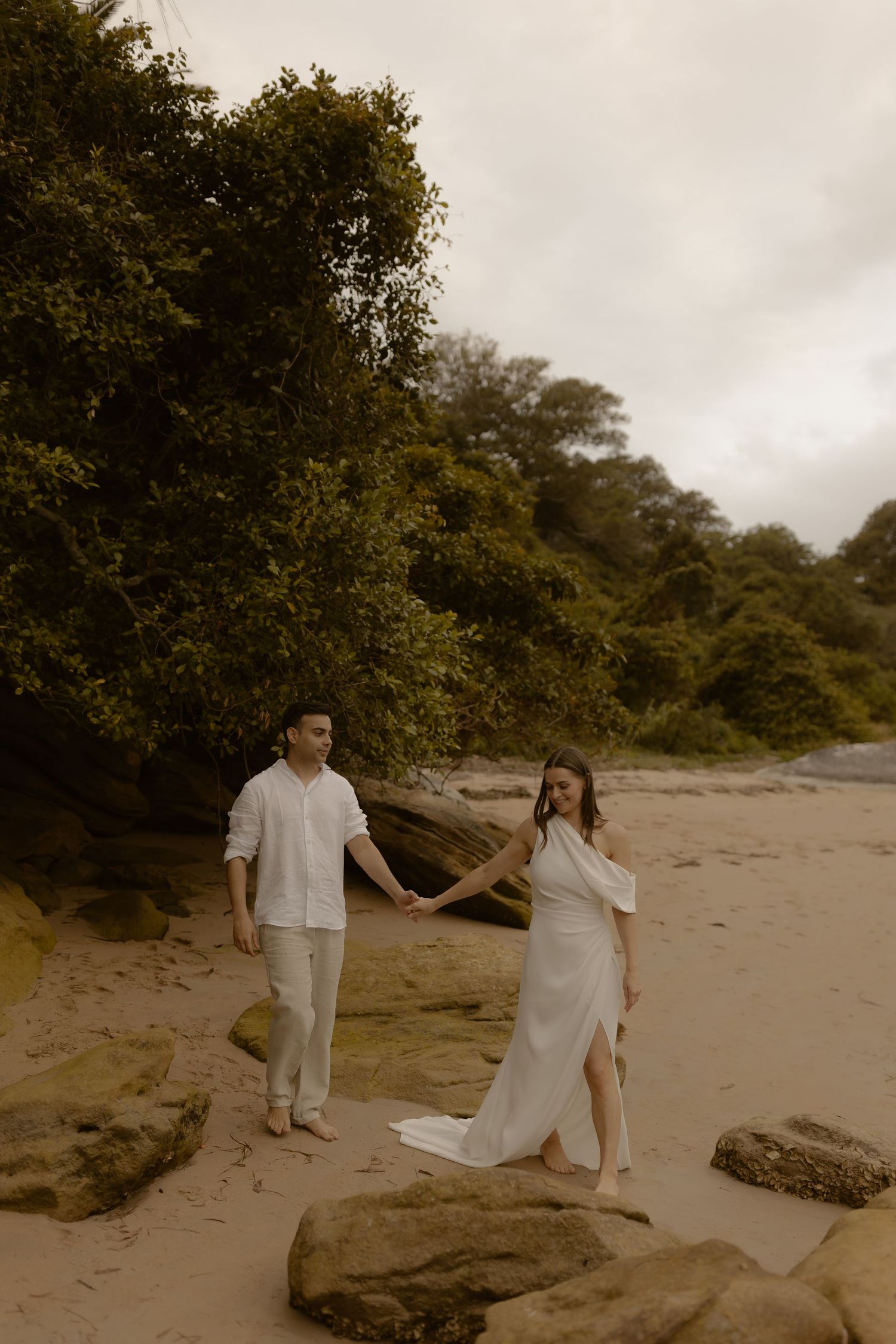 A couple dances playfully on a secluded beach with rocky cliffs and lush vegetation in the background.