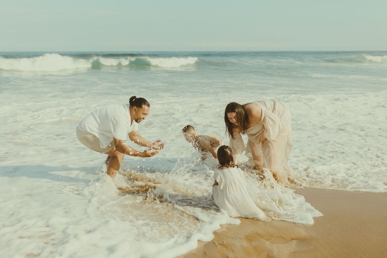 A couple in white clothing plays joyfully in the ocean waves at the beach during sunset.