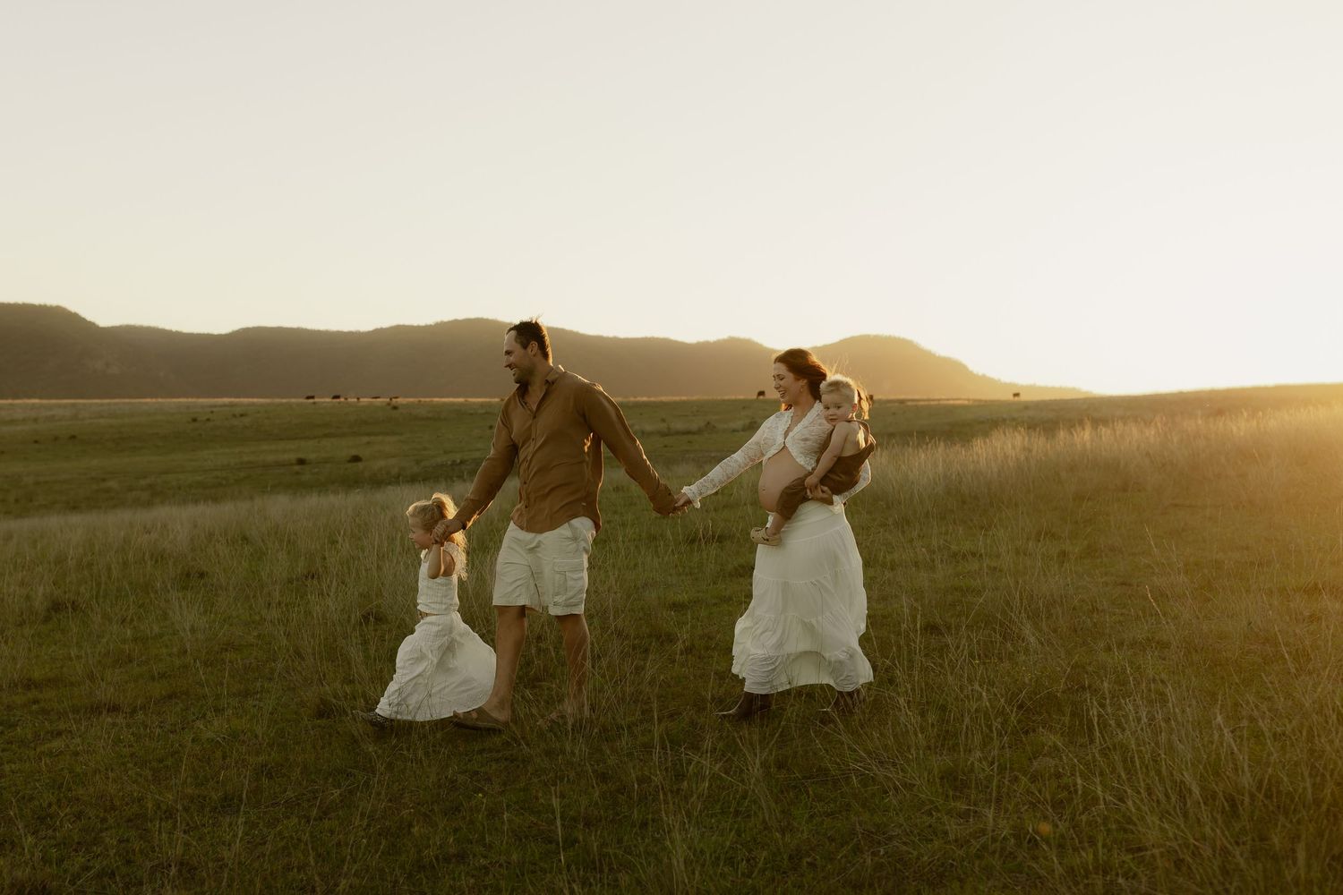 Family walking hand-in-hand through green meadow at golden sunset with mountains in background.