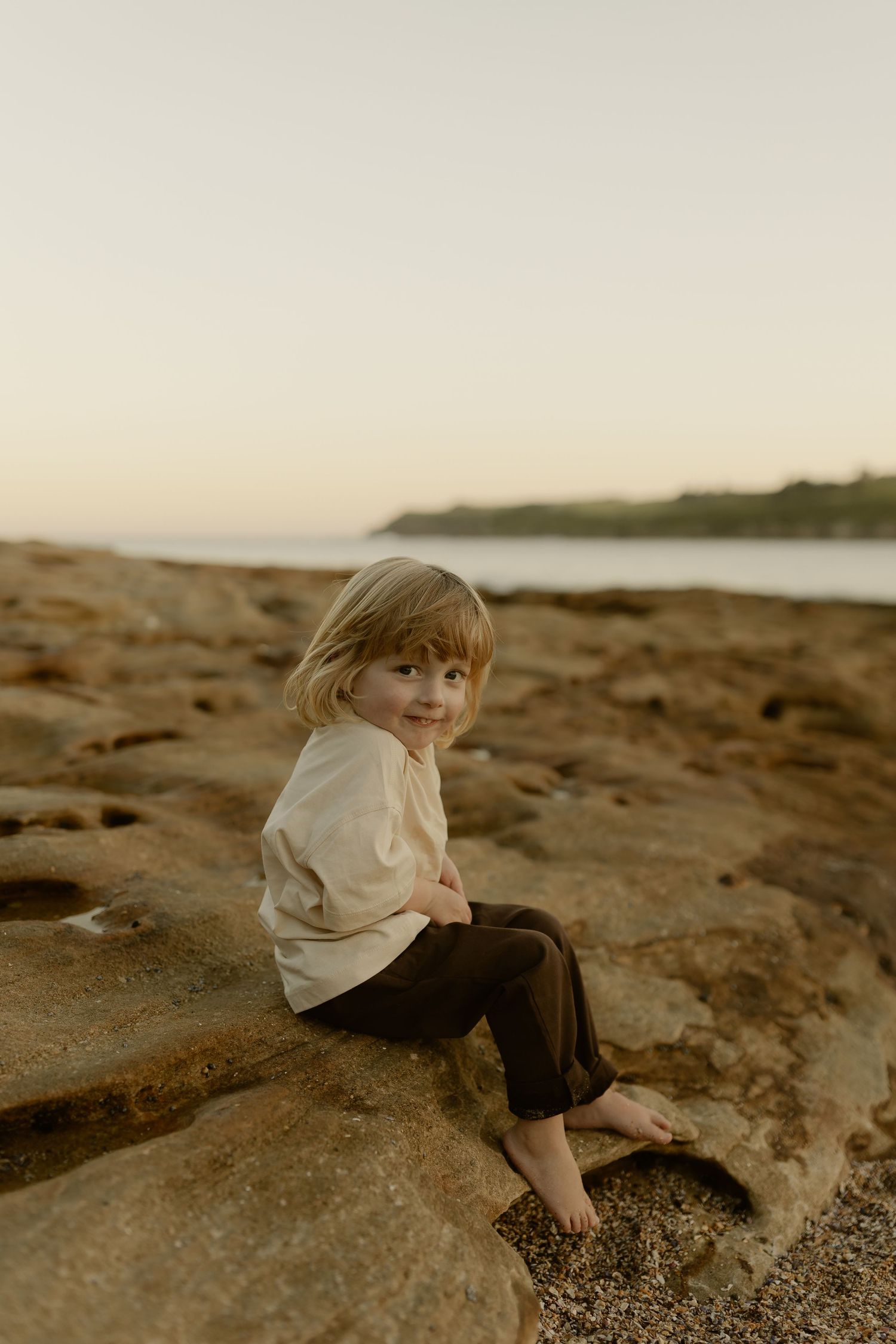 A young barefoot child with blonde hair sits smiling on rocky shoreline at sunset, wearing a cream top and dark pants.