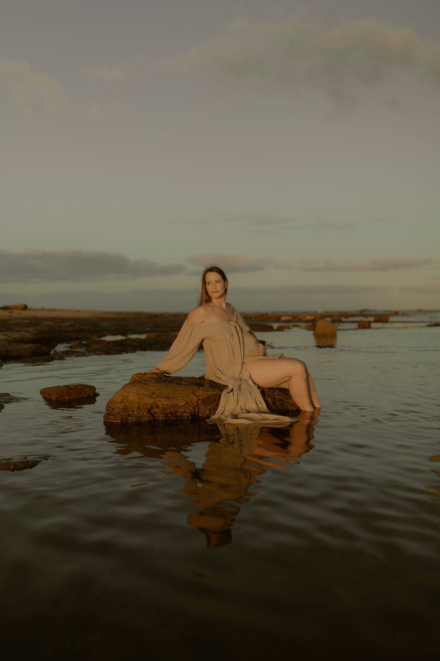 Person in light dress reclines on rocky outcrop by tranquil ocean at dusk.