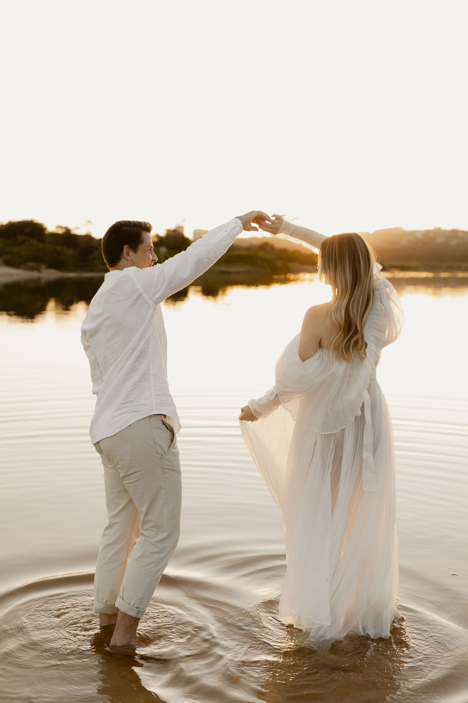 A couple in white outfits dances together in shallow water at sunset, creating ripples across the tranquil lake.