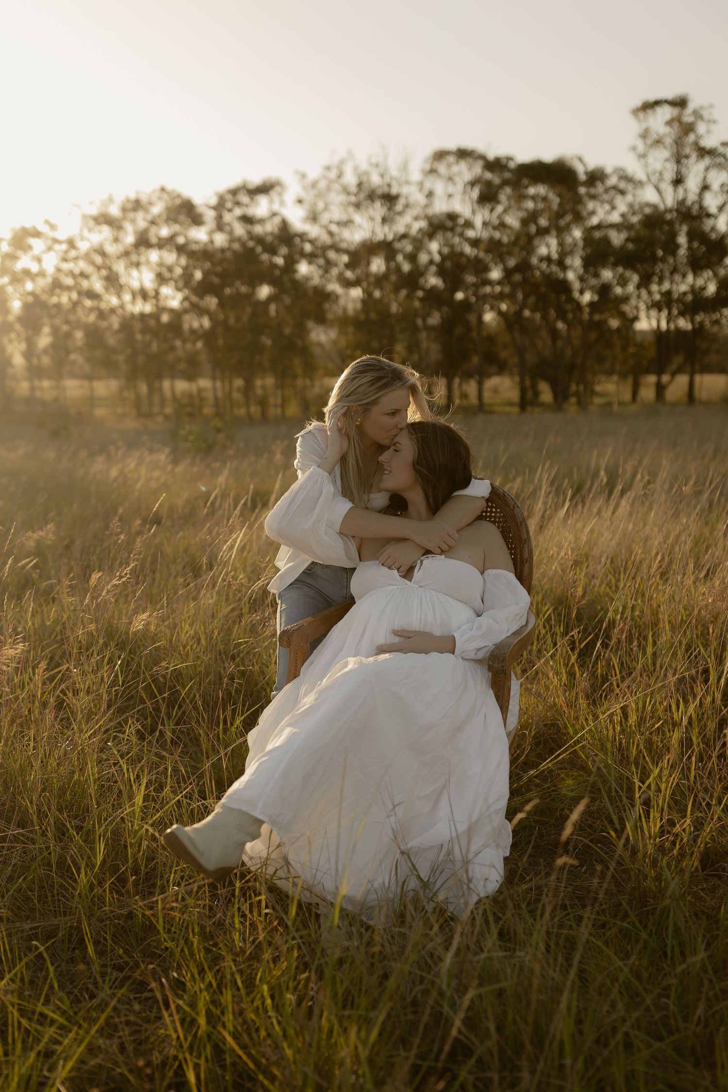 Two people in white dresses embrace in a golden field during sunset.